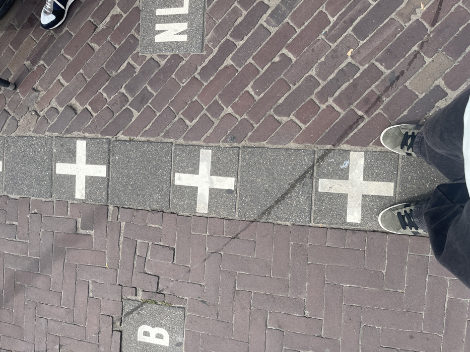 The image depicts an overhead view of a paved ground surface in Baarle-Nassau, Netherlands, illustrating the international border between Belgium (B) and the Netherlands (NL). The border is demarcated by a continuous line of rectangular grey paving slabs, each featuring a prominent white cross.
To the left of this border line, representing Belgium, the ground is paved with reddish-brown bricks laid in a herringbone pattern. A grey square tile with a white capital "B" is embedded within this paving.
To the right of the border line, representing the Netherlands, the ground is paved with reddish-brown bricks laid in a diagonal pattern. A grey square tile with white capital letters "NL" is embedded within this paving.
A person is positioned at the bottom center of the frame, with their feet straddling the international border. This individual wears dark grey suede sneakers with black laces and dark-colored pants. Their left foot is on the Belgian side, and their right foot is on the Dutch side. In the upper right corner, another person's right foot, wearing a dark blue and white sneaker, is partially visible on the Dutch side of the paving. Sunlight casts distinct shadows across the ground surface.