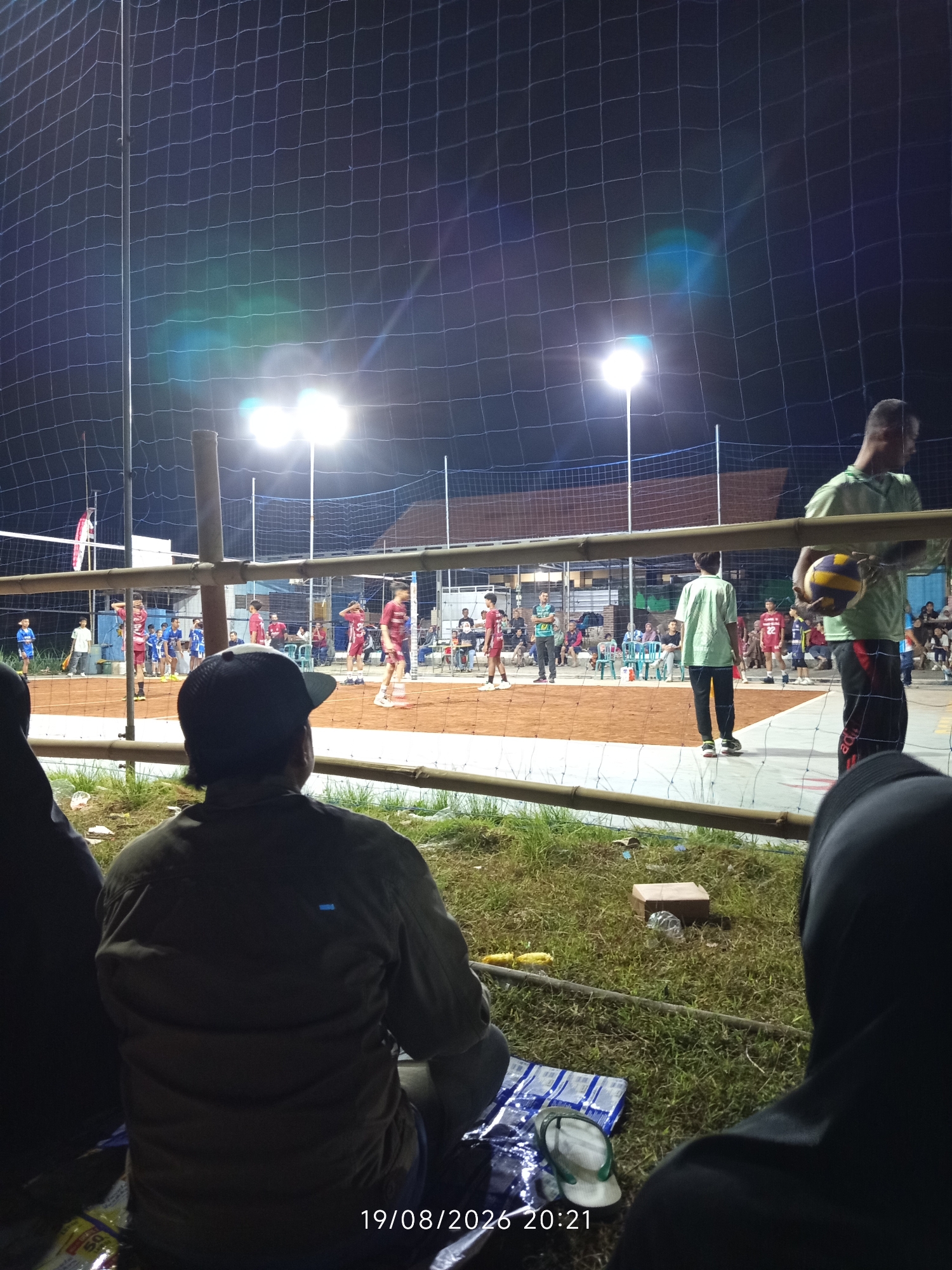 The image depicts a volleyball match at night in Canggu, Indonesia, illuminated by bright stadium lights. The scene shows a clay court with players in red and blue uniforms actively engaged in the game. Spectators, some seated and others standing, are visible behind a net, observing the match. The foreground features the backs of two individuals, one wearing a dark cap and the other a black headscarf, suggesting they are among the audience. The environment appears to be an outdoor court with grass surrounding the playing area. A date stamp at the bottom of the image indicates "19/08/2026 20:21".