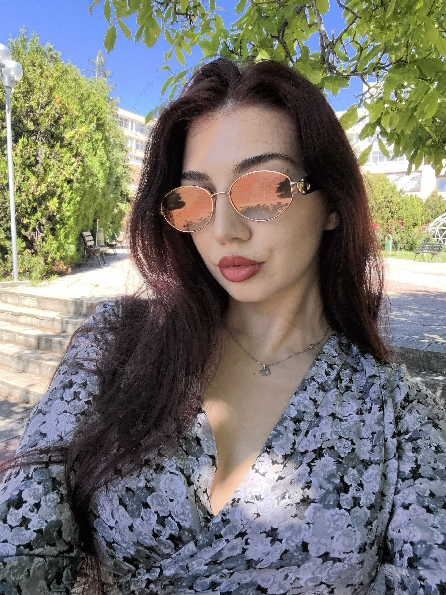A medium shot depicts a woman outside, likely in a park or garden. She wears sunglasses with reflective lenses, revealing a partial view of the surrounding environment. Her dress is floral-patterned, with a low neckline, and a silver necklace with a heart pendant is visible. In the background, there are trees, a building, benches, and a lamp post, indicating an outdoor, public area. The location is specified as the city of Unknown, Unknown.