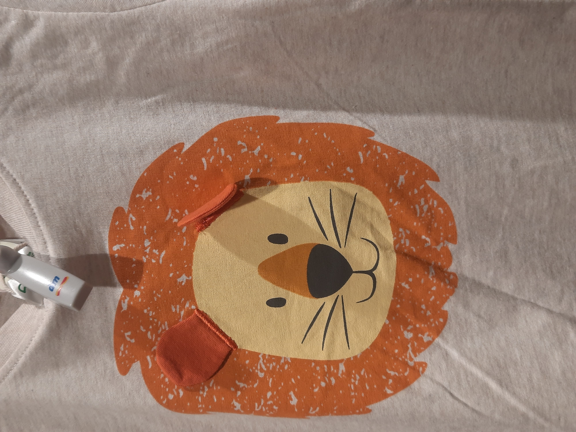 The image shows a close-up of a light-colored t-shirt with a graphic of a lion's face. The lion's face is primarily orange with a yellow circular center for its face. It has black oval eyes, a black nose, and a simple black smiling mouth. There are black whiskers extending from its nose. The lion's mane is depicted as textured orange fur. Attached to the top of the t-shirt, near the collar, is a white tag with the blue letters "dm" and a small multicolored logo, likely a brand tag. The t-shirt fabric is visible, showing a ribbed texture. The image is taken from a close-up perspective, focusing on the t-shirt graphic and the tag. There is no visible indication of the city of Rosenheim, Germany in the image.