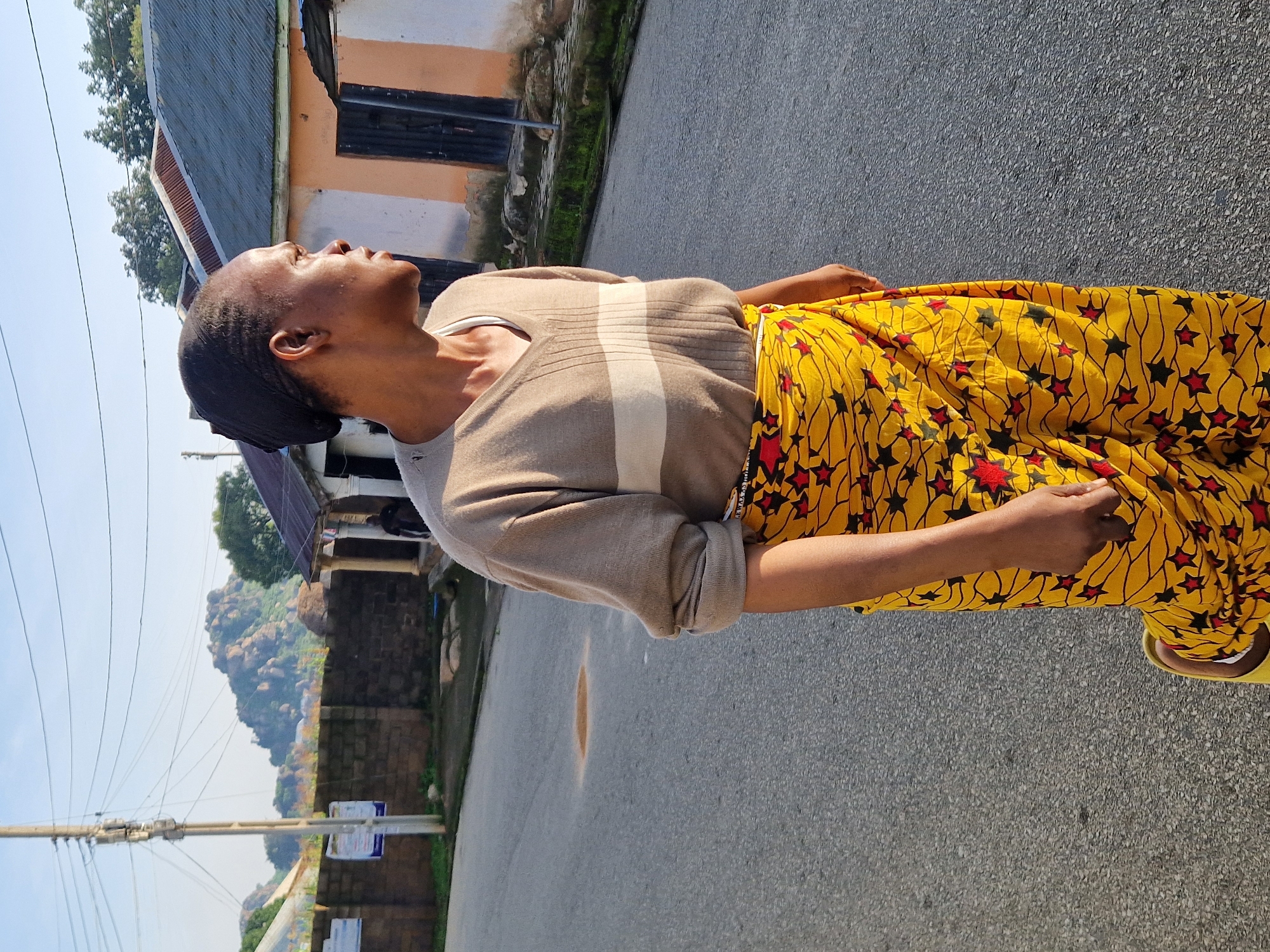 A person, identified as a woman by her attire, stands on an asphalt road in what appears to be a rural or semi-urban area. She is looking to her right, away from the camera. She wears a light brown V-neck sweater with horizontal stripes of a lighter shade, and a vibrant yellow wrap skirt adorned with a pattern of red and black stars. Her right hand is placed inside her skirt. On her feet, she wears yellow sandals.
The road is bordered by a low, dark brick wall on the left, behind which are some buildings and vegetation. A utility pole with numerous wires stands to the left of the frame. In the background, there are buildings with corrugated metal roofs and peach-colored walls with dark doors. Further in the distance, rocky hills are visible under a clear, light blue sky. The lighting suggests it is either early morning or late afternoon, with the sun casting a warm glow. A small, circular patch of discoloration is visible on the asphalt in front of the woman.