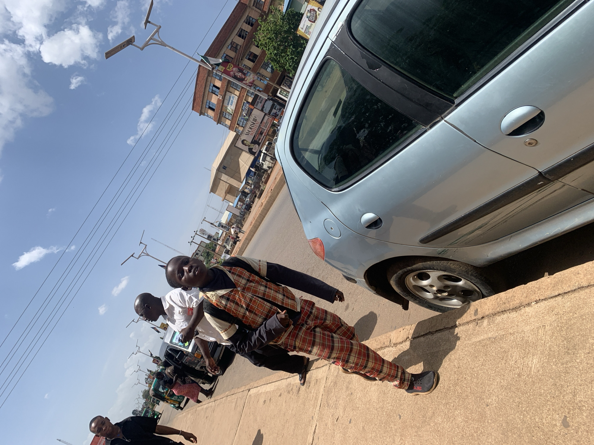 The image presents a sunlit street scene in Jos, Nigeria, featuring several individuals and vehicles. The sky is bright blue with scattered white clouds, and strong shadows indicate it is midday or early afternoon.
In the foreground, a young man is walking on a concrete sidewalk towards the viewer. He is wearing a distinctive ensemble of an orange, red, black, and white plaid long-sleeved shirt over a black underlayer, paired with matching plaid trousers and black sneakers with red accents. He looks directly at the camera with a slight smile. Adjacent to him, a light blue hatchback car, possibly a Peugeot 206, is parked roadside. Its rear tire, passenger side door, and handle are clearly visible.
Behind the primary subject, another man in a white shirt and black trousers is seen, holding a reflective object. Further along the street, other pedestrians are visible, along with a number of tricycles (keke napep) and cars on the paved road. Modern streetlights with dual fixtures, possibly solar-powered, line the street.
In the background, multi-story commercial buildings rise, adorned with various billboards and signs. Partially legible text on these signs includes "MANSURI," "360," "WORSHIP WITH US," "GOOD LIFE," and "DIVINE." Power lines are also visible stretching across the sky. The scene depicts a typical bustling urban environment.