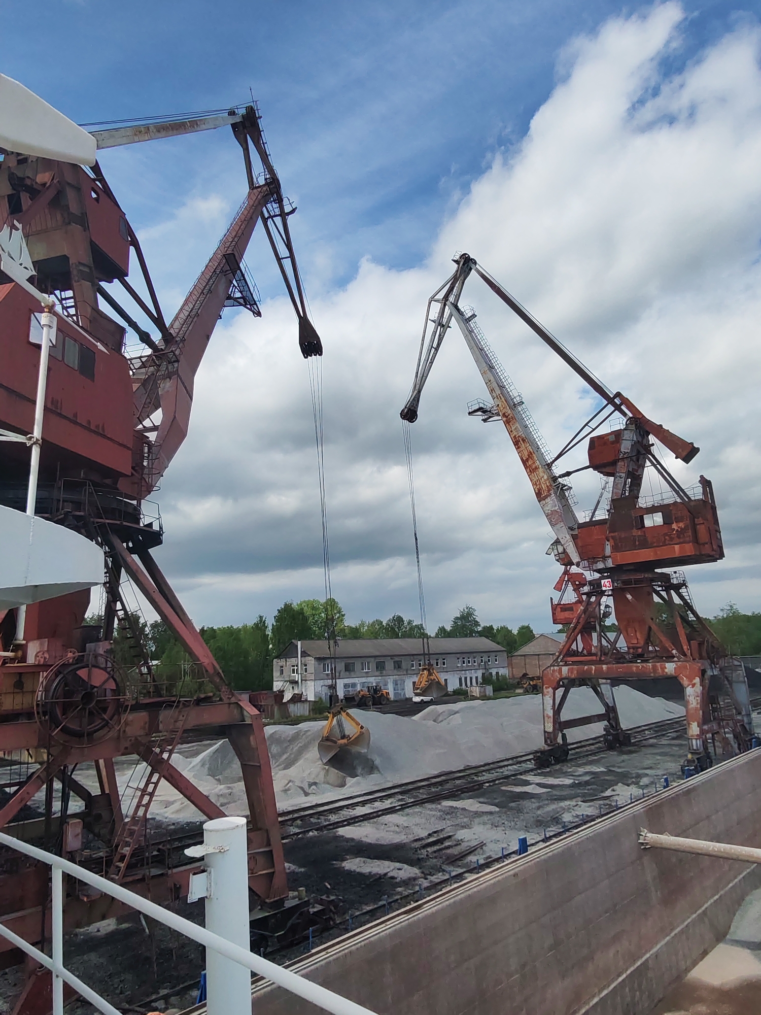 This image depicts an industrial port or loading facility in Medvezhyegorsk, Russia, under a partly cloudy sky with patches of blue. The scene is dominated by two large, weathered, red gantry or portal cranes, both showing significant rust and signs of heavy use.
The crane on the right, bearing the number "43" on its cabin, is positioned over a large pile of light-colored aggregate, with its grab bucket suspended nearby, suggesting active loading or unloading operations. The crane on the left is also oriented as if ready for operation.
In the foreground on the right, the flat-sided hull of a large vessel, likely a barge or bulk carrier, is visible, indicating that materials are being transferred to or from it. Between the cranes and the background buildings, multiple rail tracks run through the facility, flanked by substantial piles of both light-grey aggregate (possibly sand or gravel) and darker, coarser material (potentially coal or crushed stone).
Several utilitarian, multi-story industrial buildings are visible in the background, partially obscured by green trees. The overall setting is one of heavy industry and bulk material handling. No people are visible in the image. The time of day appears to be daytime, with ample natural light.