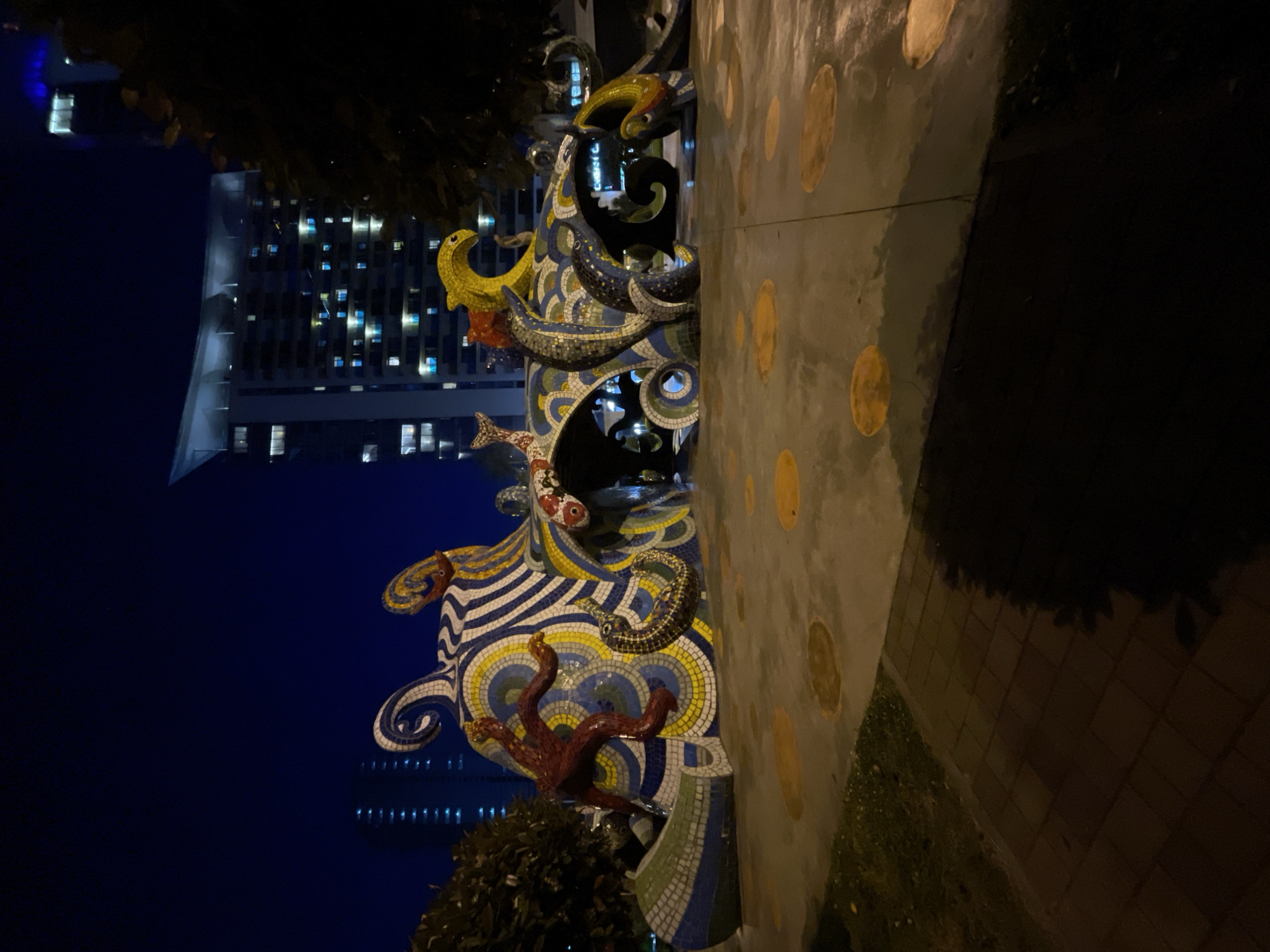 The image captures a nighttime scene in Batumi, Georgia, focusing on a large, vertical mosaic sculpture. The sculpture appears to be themed around marine life, featuring various mosaic renderings of fish, sea creatures, and waves in a vibrant array of colors including yellow, blue, white, and red. The base of the sculpture is partially obscured by foliage.
In the background, a modern high-rise building is visible with numerous lit windows, contrasting against the deep blue night sky. A concrete wall with evenly spaced, circular indentations runs alongside the sculpture, marking the edge of a walkway. The right side of the image fades into darkness, suggesting a poorly lit area. The perspective is from a slightly low angle, emphasizing the height of the sculpture and the building behind it. The image is rotated for some reason.