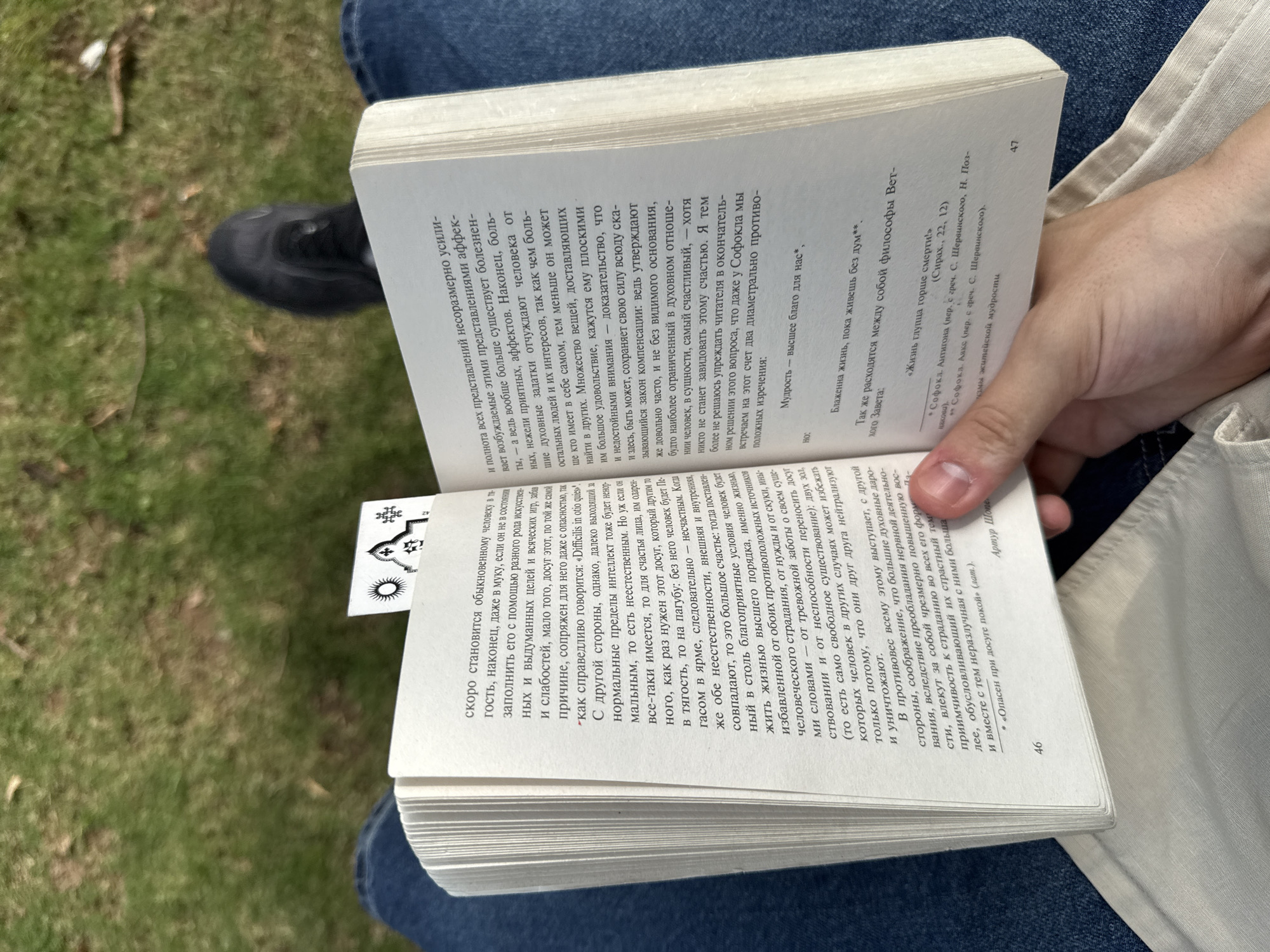 A person sits outdoors with a book open in their hands, the left page marked with a stylized bookmark featuring a sun and other symbols. The right page has text in Russian, including quotes from Sophocles and references to "wisdom." The pages are numbered 46 and 47, indicating the book is in progress. The person is wearing blue jeans and a light-colored garment. A black shoe is visible in the background, and greenery is sparsely seen. No indication of Hà Nội is present.