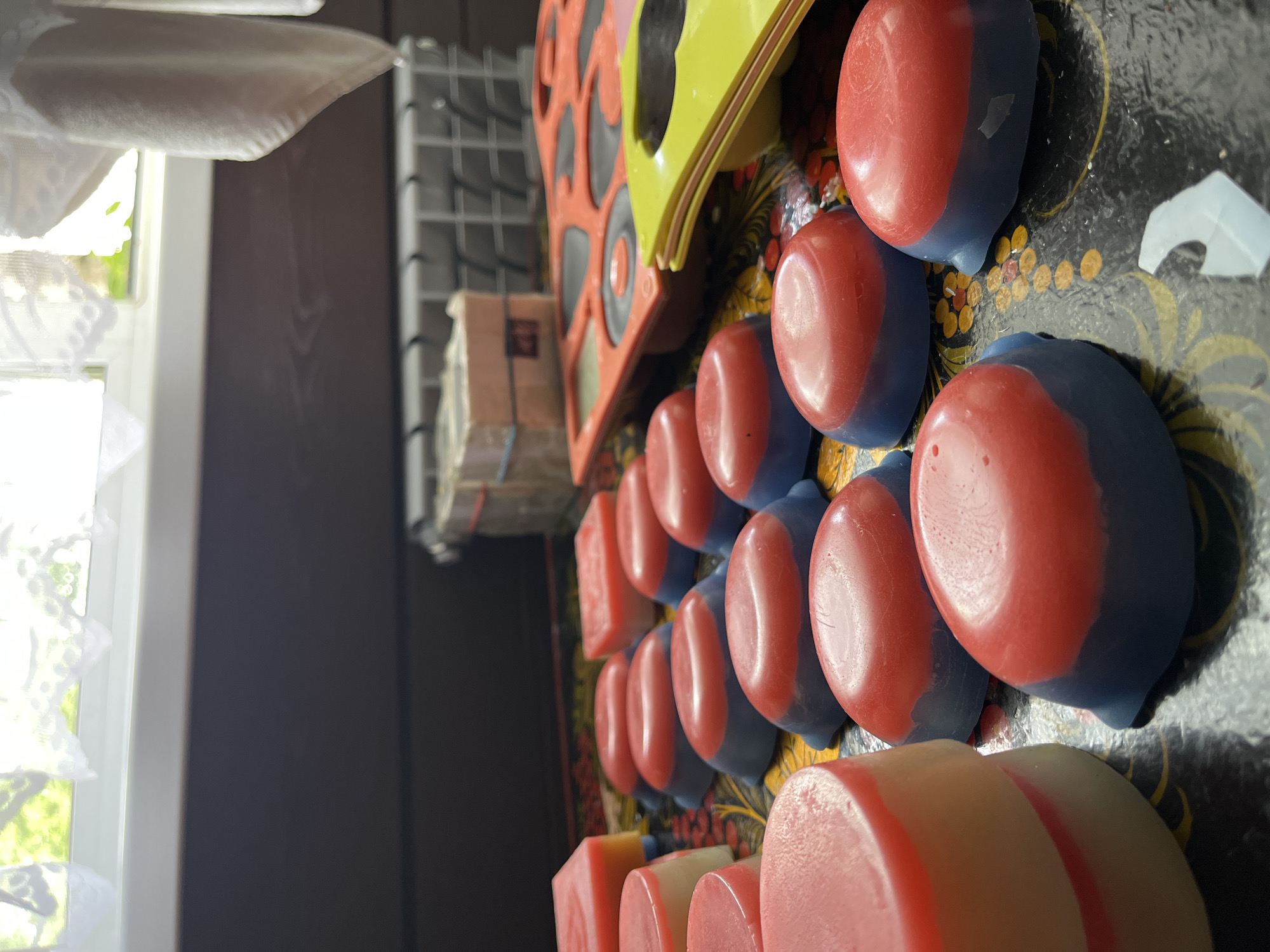 The image depicts a collection of handmade soaps arranged on a decorative tray. The soaps are predominantly red and blue, with some variations in shape and size. In the background, a window with white lace curtains is visible, allowing natural light to illuminate the scene. Below the window, a dark wooden wall and a radiator are present. A red silicone mold and a yellow plastic mold are also visible, suggesting that these soaps were recently made or are in the process of being made. There are no people present in the image. The scene appears to be indoors, likely in a domestic setting. There is no information to indicate this location is in Divnogorsk, Russia.