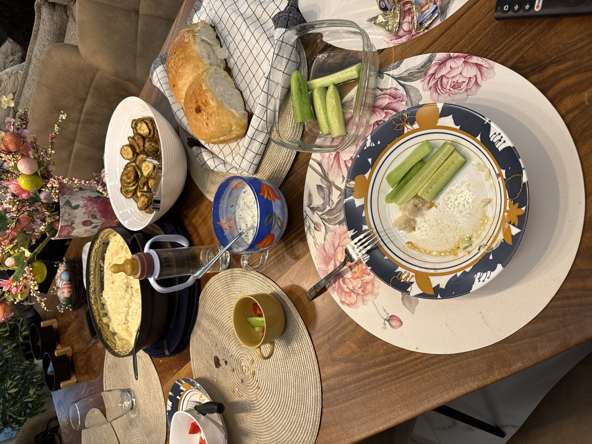 The image shows a table in what appears to be a home, laden with food. The table is wooden and covered with placemats. There's a bowl of fried zucchini, bread in a cloth-lined basket, and a glass container with cut cucumbers. A blue patterned mug containing a white sauce is present, along with a small yellow cup with tomato pieces and sliced cucumber inside. There is a plate with remnants of food and cucumber slices, accompanied by a fork. A pot, possibly containing fondue, stands next to a bottle propped up by a white holder. A vase of flowers with decorated Easter eggs is visible. The environment is domestic and cluttered, suggestive of a casual dining setting. The location is not explicitly mentioned, but the prompt asks us to assume it is in Sofia, Bulgaria. The image lacks people; it's focused on the food arrangement.