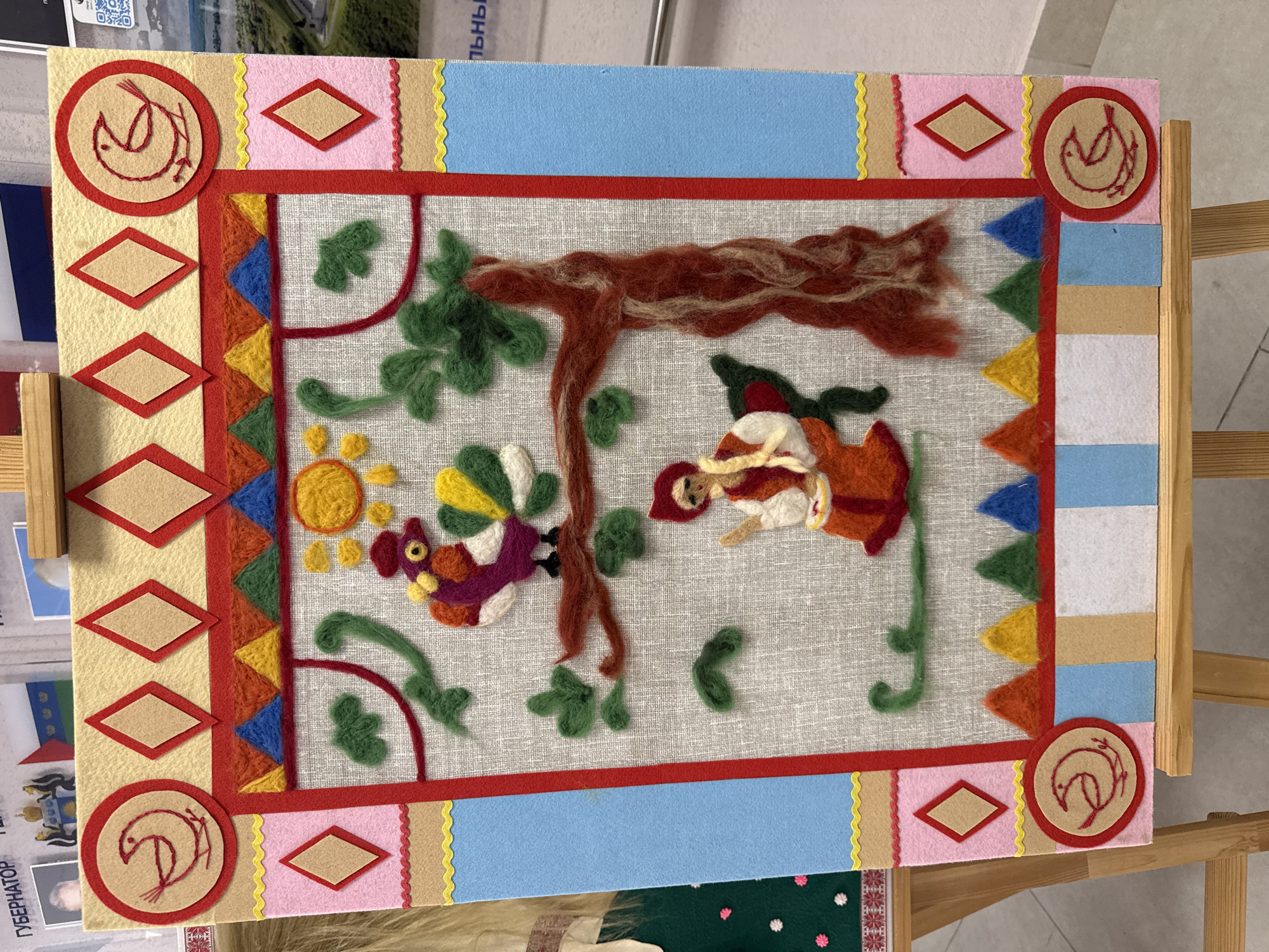 The image shows a colorful felt and yarn artwork, likely handmade. The artwork depicts a scene with a tree, a sun, a bird, and a woman. The woman is wearing a red dress and is possibly spinning or weaving thread. The bird has colorful plumage. The background is a light gray fabric, and the artwork is framed by layers of felt in various colors, including red, blue, pink, and yellow. The frame also includes geometric shapes like diamonds and circles. The artwork seems to be resting on a wooden easel or stand. Based on the context in the surrounding images, the artwork is photographed in Tobolsk, Russia.
