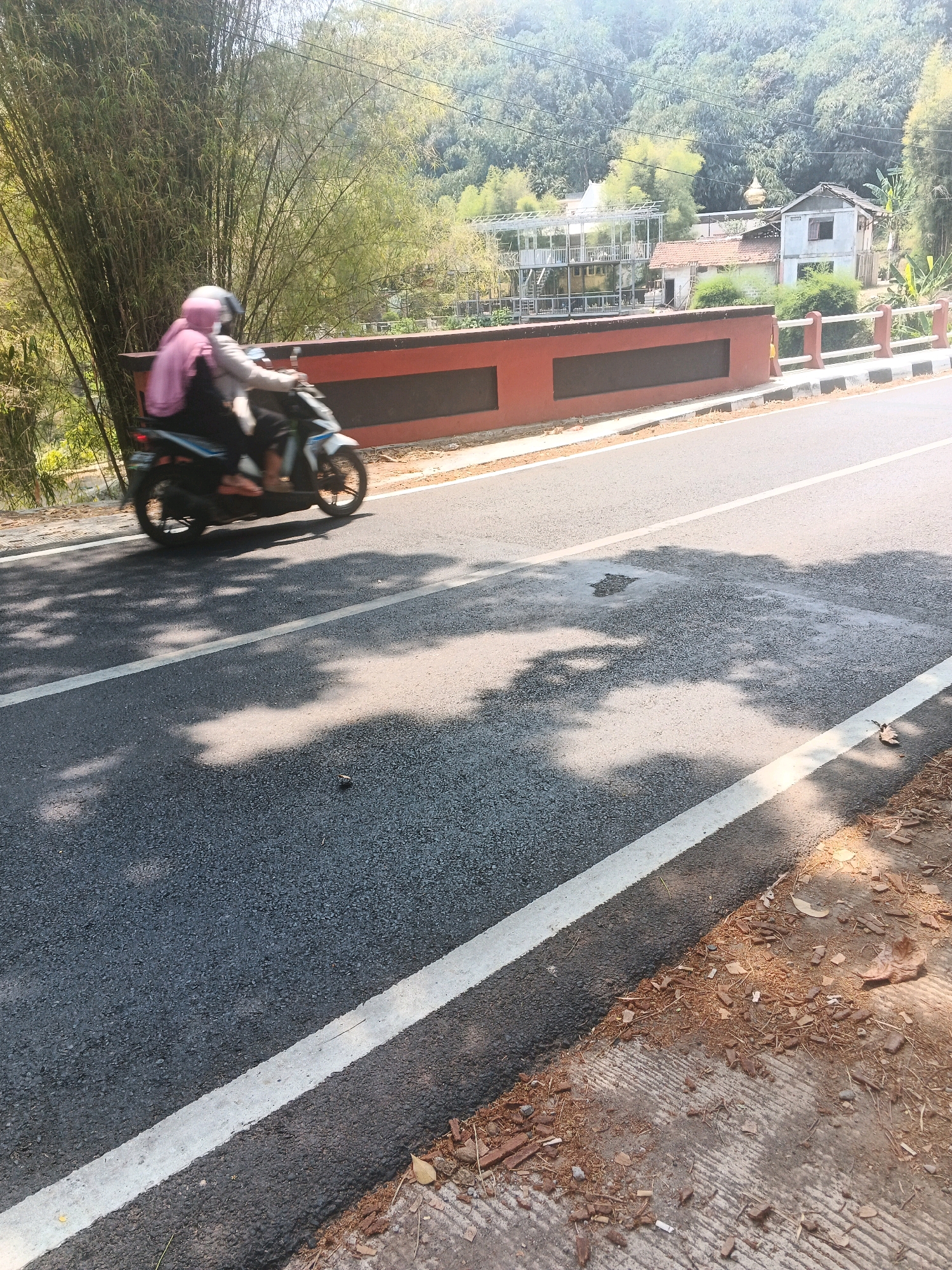The image depicts a street scene in Semarang, Indonesia, with a view of a bridge and a forested hillside. In the foreground, two people are riding a motorcycle on an asphalt road with a white dividing line. The rider and passenger are wearing helmets, and the passenger is wearing a pink hijab. Bamboo trees are visible on the left side of the road, and a red-colored bridge railing runs along the side of the road. In the background, several buildings, including houses and a structure with a metal frame, are situated on the hillside, surrounded by lush green trees. The sun is shining, casting shadows on the road.