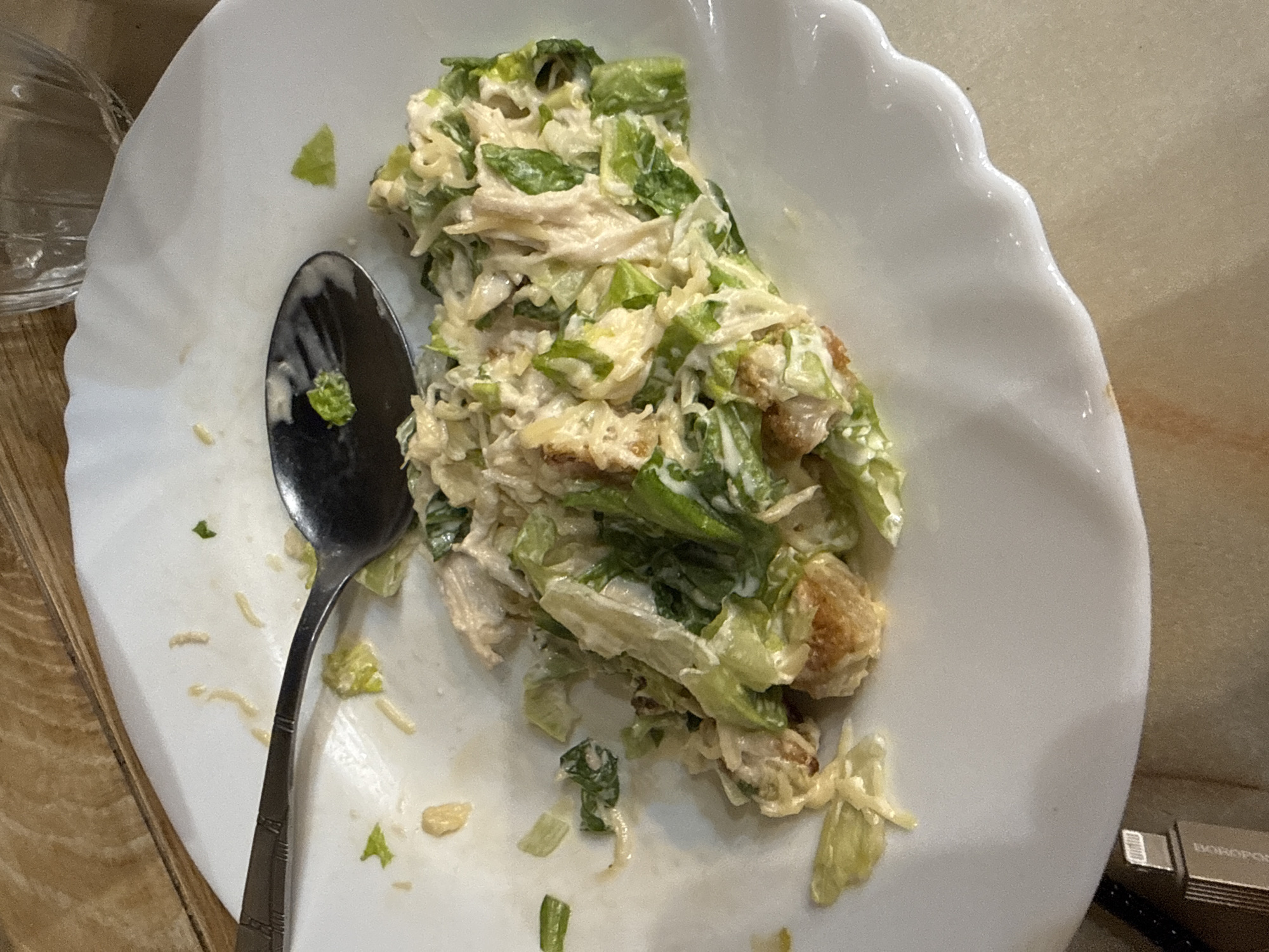 The image displays a close-up, overhead view of a partially eaten Caesar salad with chicken, served on a white plate in Birobidzhan, Russia. The salad consists of chopped romaine lettuce, shredded chicken, and a creamy white dressing. Crispy, golden-brown croutons are visible throughout the salad, alongside fine strands of what appears to be grated Parmesan cheese.
A silver-colored spoon with decorative etchings on its handle rests on the left side of the plate, partially immersed in the salad. Around the main portion of the salad and beneath the spoon, there are scattered pieces of lettuce, drops of dressing, and cheese, indicating that some of the meal has already been consumed.
The white plate features a distinctive scalloped edge along its right side. The setting appears to be indoors, with a light-colored wooden surface visible as a table in the background on the left and bottom left. A clear glass is partially visible on the far left of the frame. On the far right, a silver USB drive is present, with the word "BOROPOP" partially legible on its side. The lighting suggests an indoor environment.