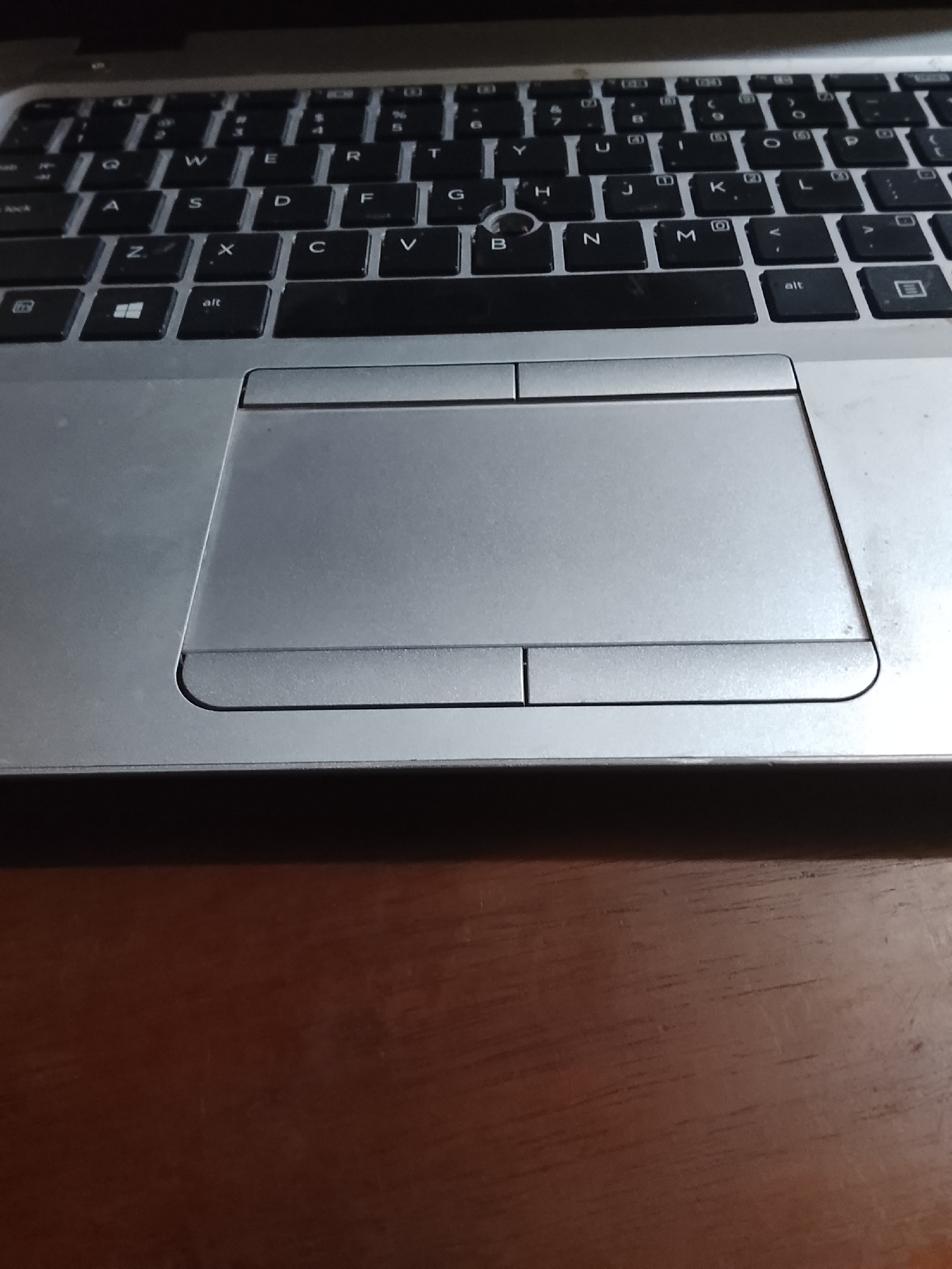 A close-up shot focuses on a silver laptop's keyboard and touchpad. The keyboard is slightly dusty, with black keys and white lettering. The touchpad is rectangular and smooth, with distinct left and right click buttons at the bottom. The laptop sits on a brown, wood-grain surface. The scene is indoors. No specific location in Multan, Pakistan, is discernible from this image. No people, actions, or interactions are captured in the frame.