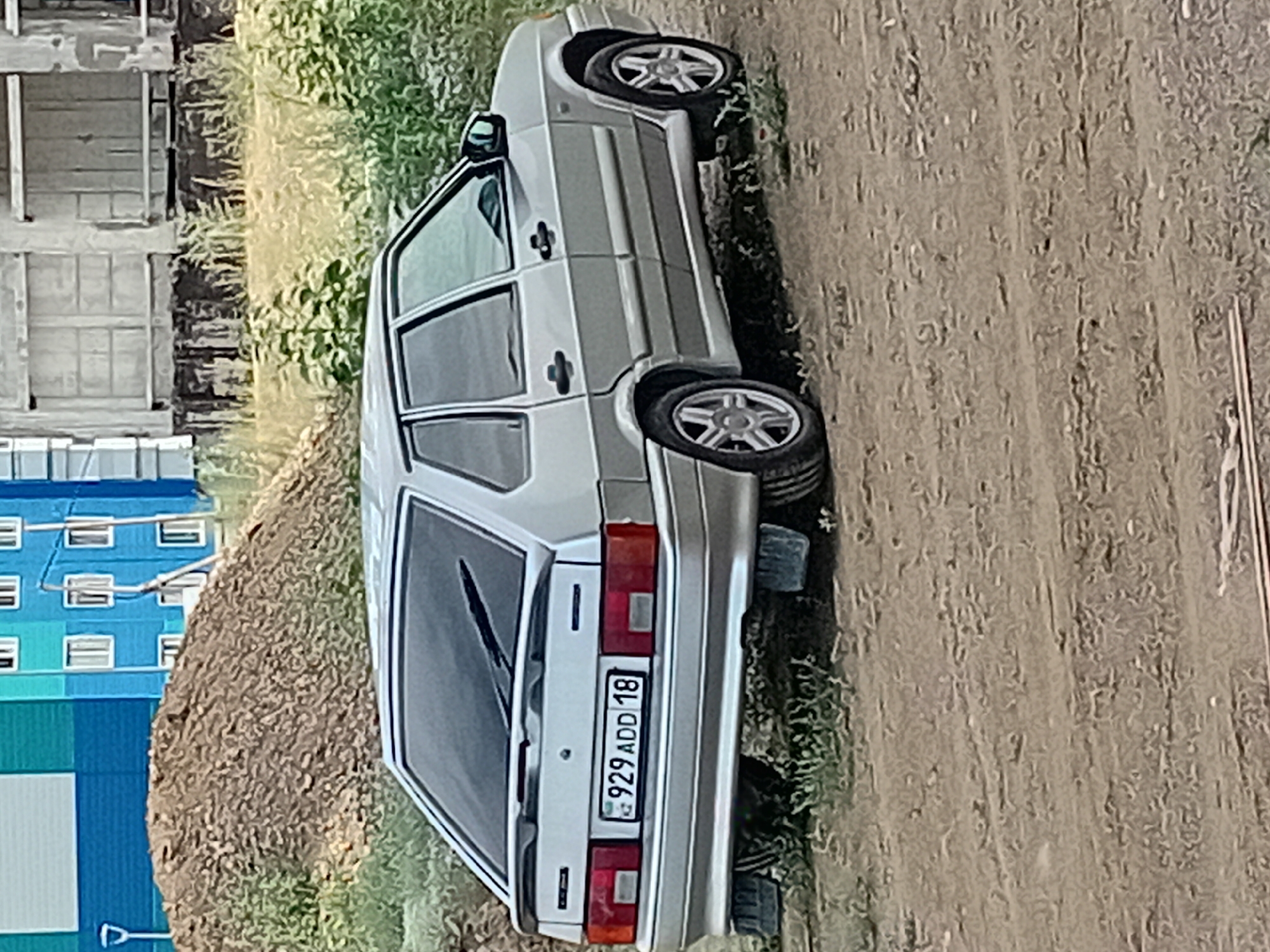 A silver Lada hatchback is parked on a dirt surface. The car has a spoiler on the rear, and a license plate with the characters "929 ADD 18" is visible. The background shows a pile of dirt, some green vegetation, and a multi-story building with blue and grey sections. The building appears to be an apartment complex, characteristic of urban areas in Semey, Kazakhstan. The scene suggests an outdoor parking area or a residential courtyard within the city.