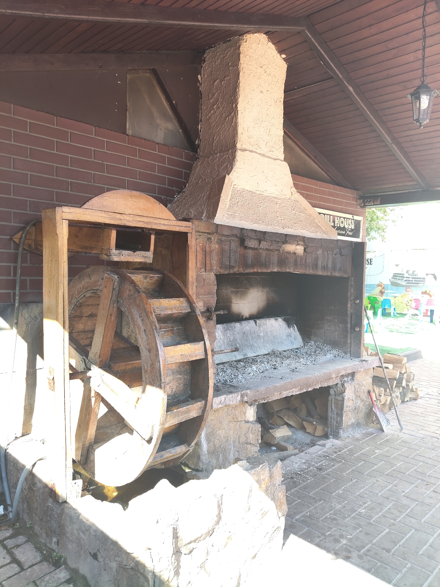 The image depicts an outdoor area featuring a large, rustic barbecue grill made of stone and brick, complete with a chimney. Adjacent to the grill is a decorative wooden water wheel, partially submerged in a stone-lined basin. The area appears to be covered by a wooden roof structure supported by columns. In the background, a brick wall is visible, along with signage that reads "GRILL HOUSE" and "American Food". Further in the background, there are colorful plastic chairs and tables set up in what looks like a seating area, with a large boat visible on the horizon. A pile of firewood is stacked next to the barbecue, and a shovel leans against the grill. The ground is paved with interlocking bricks. The scene suggests a restaurant or a recreational area designed for outdoor dining and activities. While the specific location in Constanța, Romania cannot be definitively identified from the image alone, the presence of a "Grill House" and the overall aesthetic are consistent with a commercial establishment, possibly a restaurant with an outdoor seating area. No people are clearly visible in the image.