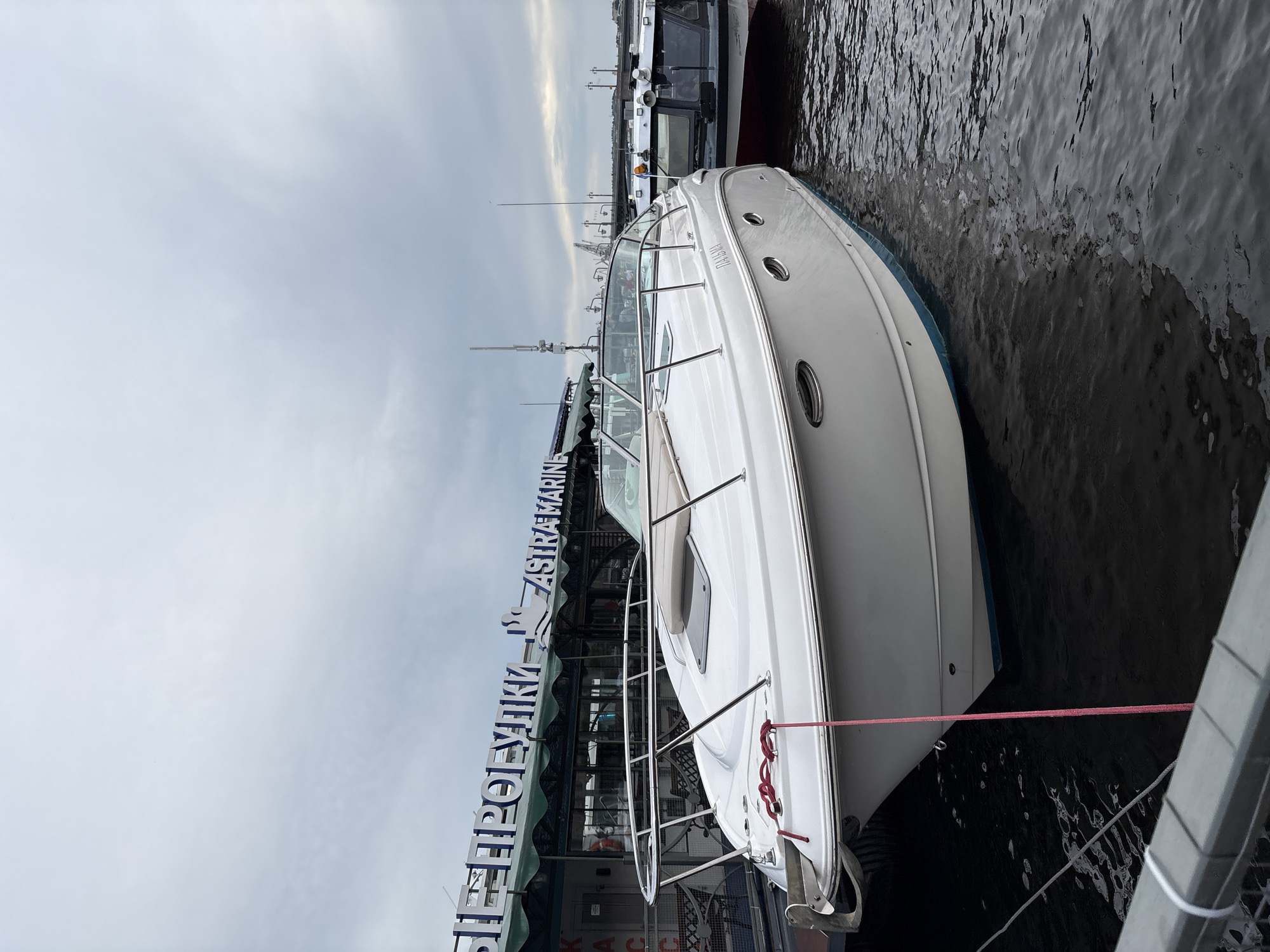 A white speedboat with a blue waterline is docked on a body of water, likely a canal or river, in Saint Petersburg, Russia, under a cloudy sky. The boat has a polished chrome railing and a windshield. A red rope is tied from the boat's bow to a dock. Behind the speedboat, another boat, dark in color with windows, is also docked. In the background, a building with large white lettering that appears to read "АСТРА МАРИН" and "ШЕПРОПЫХИ" is visible. The building has a green awning. Further in the background, a bridge and cranes suggest an urban industrial or port setting. The water is dark and rippled.