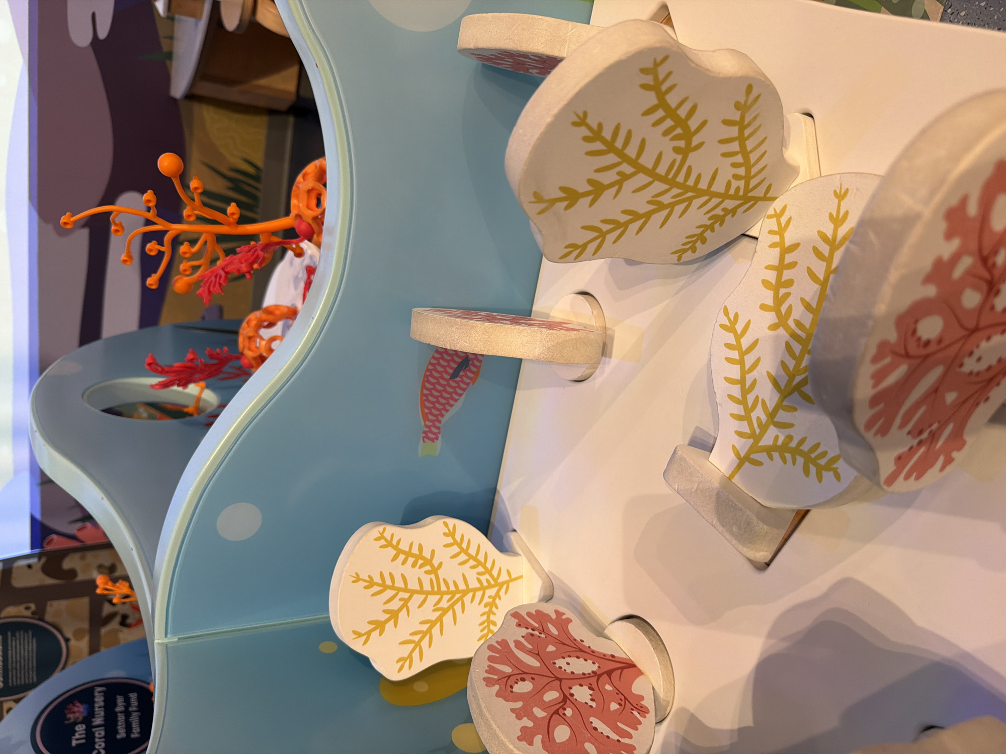 This is a close-up, eye-level, indoor photograph of a children's interactive exhibit depicting a coral nursery. The exhibit is made of molded plastic and wooden pieces, with brightly colored coral and sea creature decorations. In the foreground, several flat, wooden cutouts of coral are positioned in slots on a white surface. Two of these cutouts are printed with a pink coral pattern, and two are printed with a yellow, fern-like pattern. Behind these, a larger, curved, light blue plastic structure forms the main part of the exhibit, with cutouts for placing the coral pieces. Inside this structure, to the right, are red and orange plastic models of coral and anemones, suggesting an underwater scene. A sign with text partially visible at the top left of the image reads "The Coral Nursery" and "Setnor Byer Family Fund," indicating this is likely a museum or educational center exhibit. The lighting is bright and even, suggesting an indoor, well-lit environment. The photograph does not contain any people or animals, and there is no clear indication of the specific location within Fort Lauderdale, United States of America, beyond the general context of an interactive exhibit.
