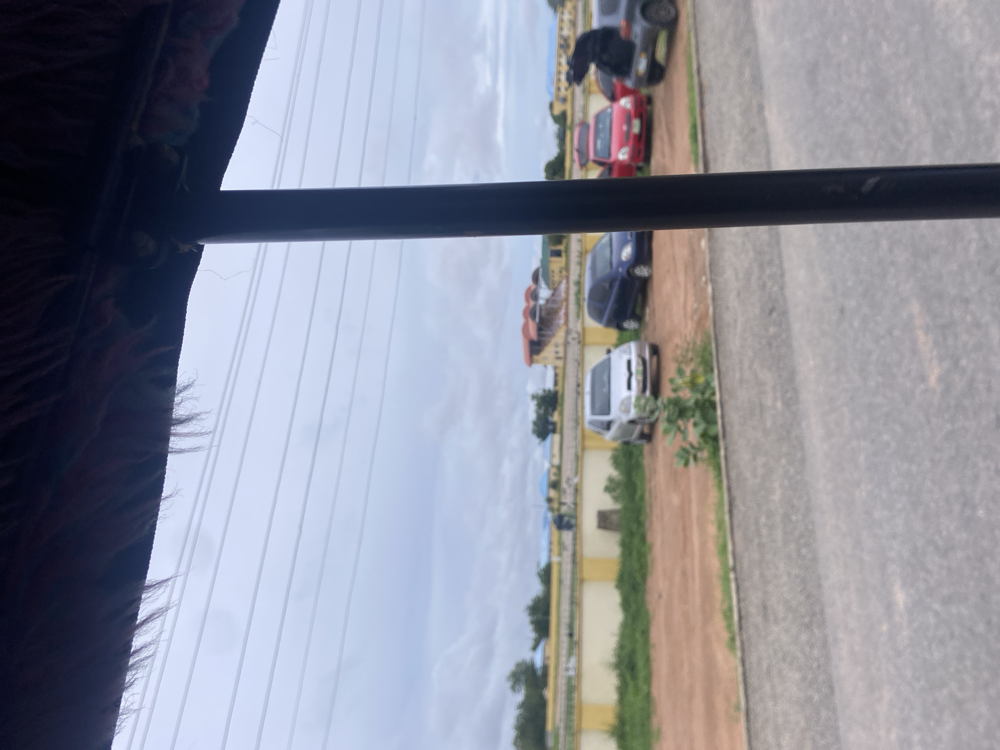 The image depicts an outdoor scene in Jalingo, Nigeria, likely on a cloudy day. In the foreground, a dark, cylindrical pole obscures part of the view, and the top of the frame shows the underside of a dark, furry structure, possibly part of a vehicle's canopy or awning. Beyond this, a line of parked cars is visible on a dirt or gravel lot. The cars include a silver hatchback, a dark blue hatchback, a red hatchback, and a dark grey SUV. The red hatchback has its rear hatch open. In the mid-ground, a low, yellow and beige wall runs horizontally across the frame, separating the parking area from a strip of green grass and vegetation. Beyond the wall, several buildings with light blue roofs are visible in the distance, along with trees. The sky is overcast with a pale, diffused light. The scene appears to be a parking area adjacent to a facility or residential area.
