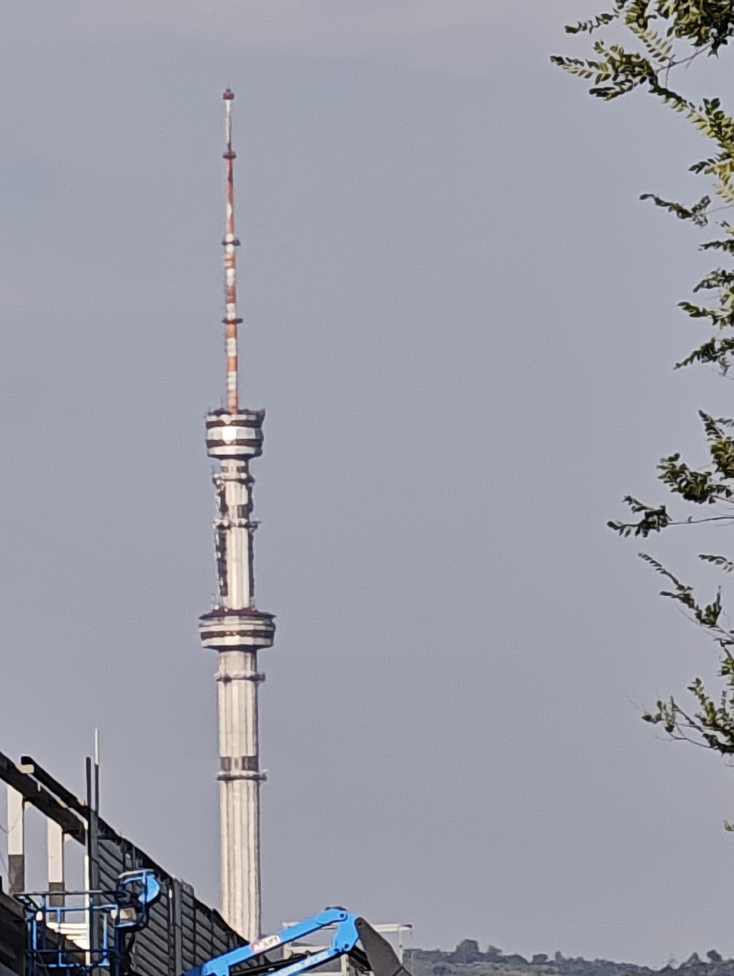 The image shows the Almaty Television Tower, also known as Kok-Tobe Tower, situated in Almaty, Kazakhstan. The tower is a tall, slender structure with distinct horizontal bands of white and red at its upper sections, transitioning to a silvery, segmented design lower down. At the base of the tower, part of a construction site is visible, including a blue aerial work platform. The sky is overcast and appears hazy. In the foreground on the right, a portion of a tree with green leaves branches into the frame.