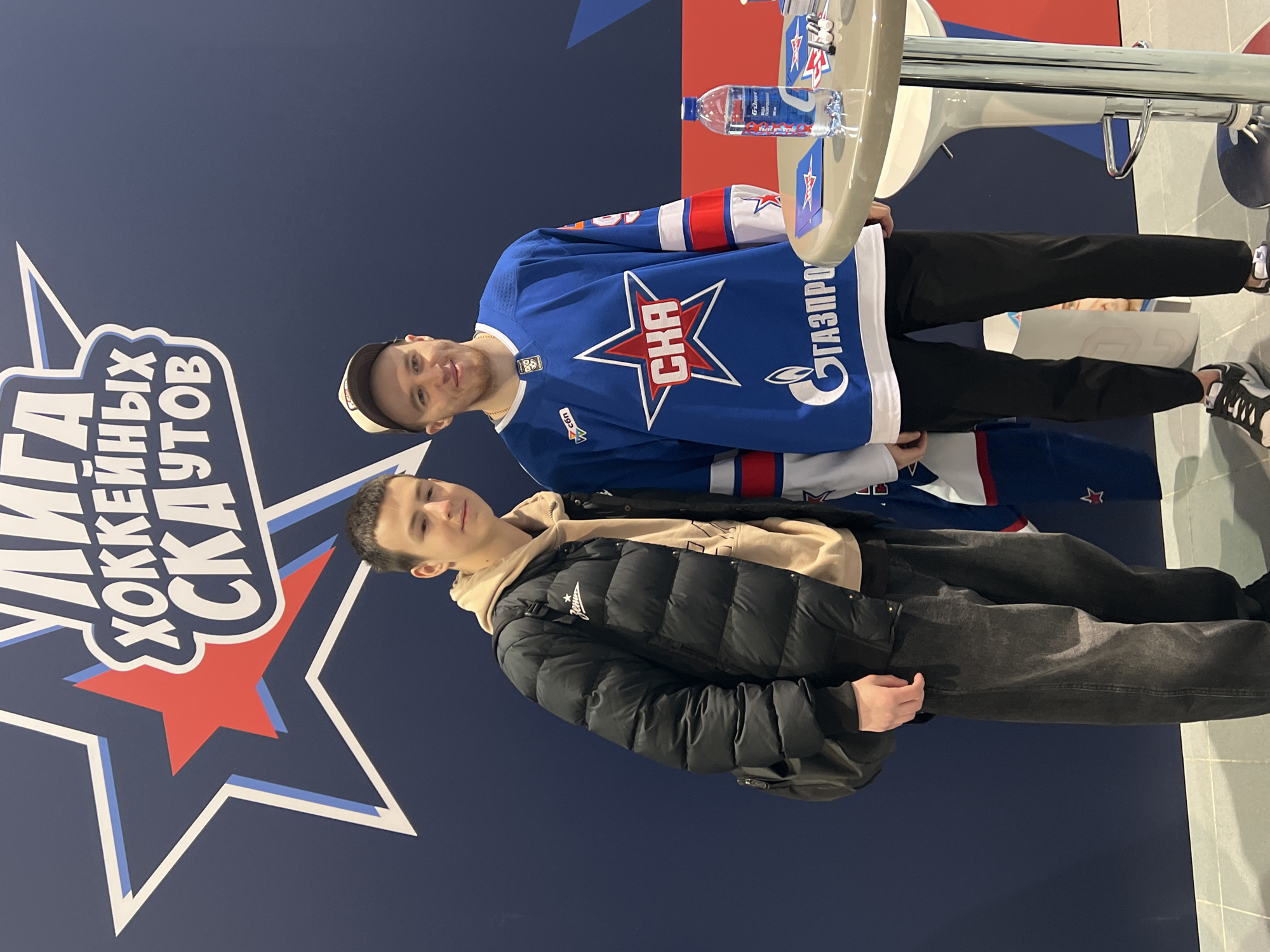 Two men stand next to each other indoors. The man on top is wearing a blue, white, and red hockey jersey with "SKA" and "ГАЗПРОМ" printed on it and a white baseball cap. The man below him is wearing a black puffer jacket over a beige hooded sweatshirt and dark gray jeans. They are standing in front of a blue wall with a large white and red star and Cyrillic text. There is a table with a water bottle on the upper right corner of the image. The city is likely Saint Petersburg, Russia, based on the hockey jersey branding.