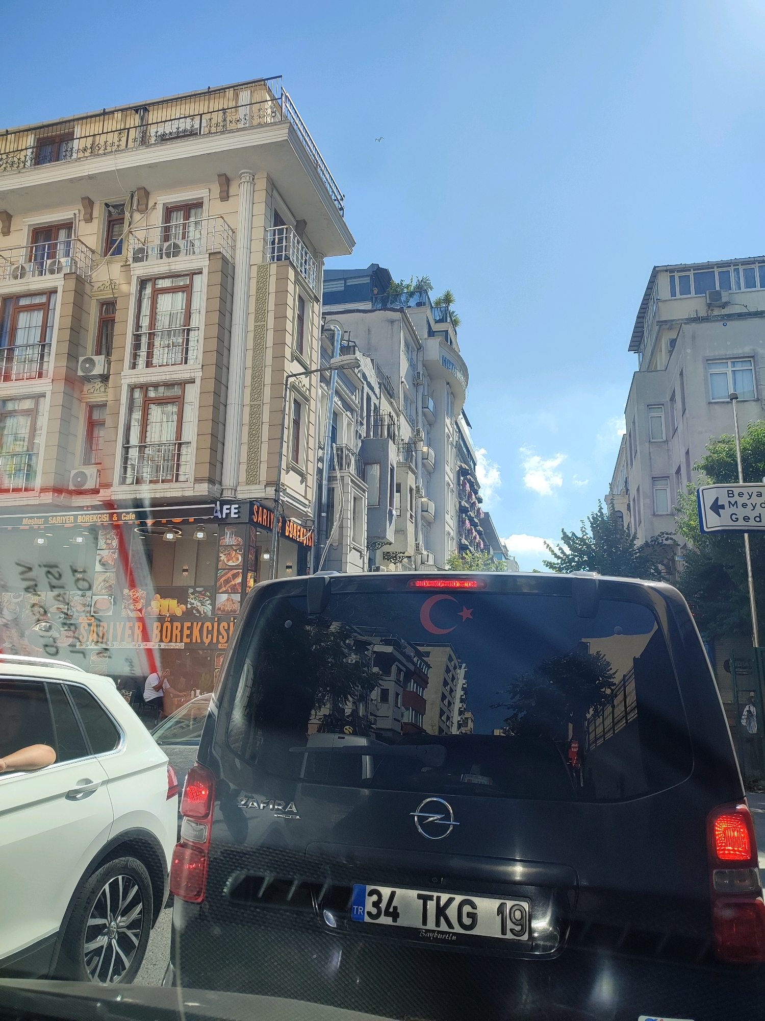 The image shows a street scene in Istanbul, Turkey. In the foreground, the rear of a dark-colored Opel Zafira minivan is visible, with its rear windshield reflecting buildings and trees. A Turkish flag, a red crescent moon and star, is affixed to the rear windshield. The license plate is visible, displaying "34 TKG 19 TR". To the left of the minivan is a white SUV. In the background, there are several multi-story buildings with balconies and windows, typical of urban architecture. One building appears to house a cafe or restaurant, with signage reading "Meshur SARIYER BÖREKÇİSi & Cafe". The sky is clear blue with some white clouds. A street sign with Turkish writing, possibly indicating directions to "Beyazit Meydanı" and "Gedikpaşa", is visible on the right side of the image. The scene depicts a street in Istanbul on a sunny day.