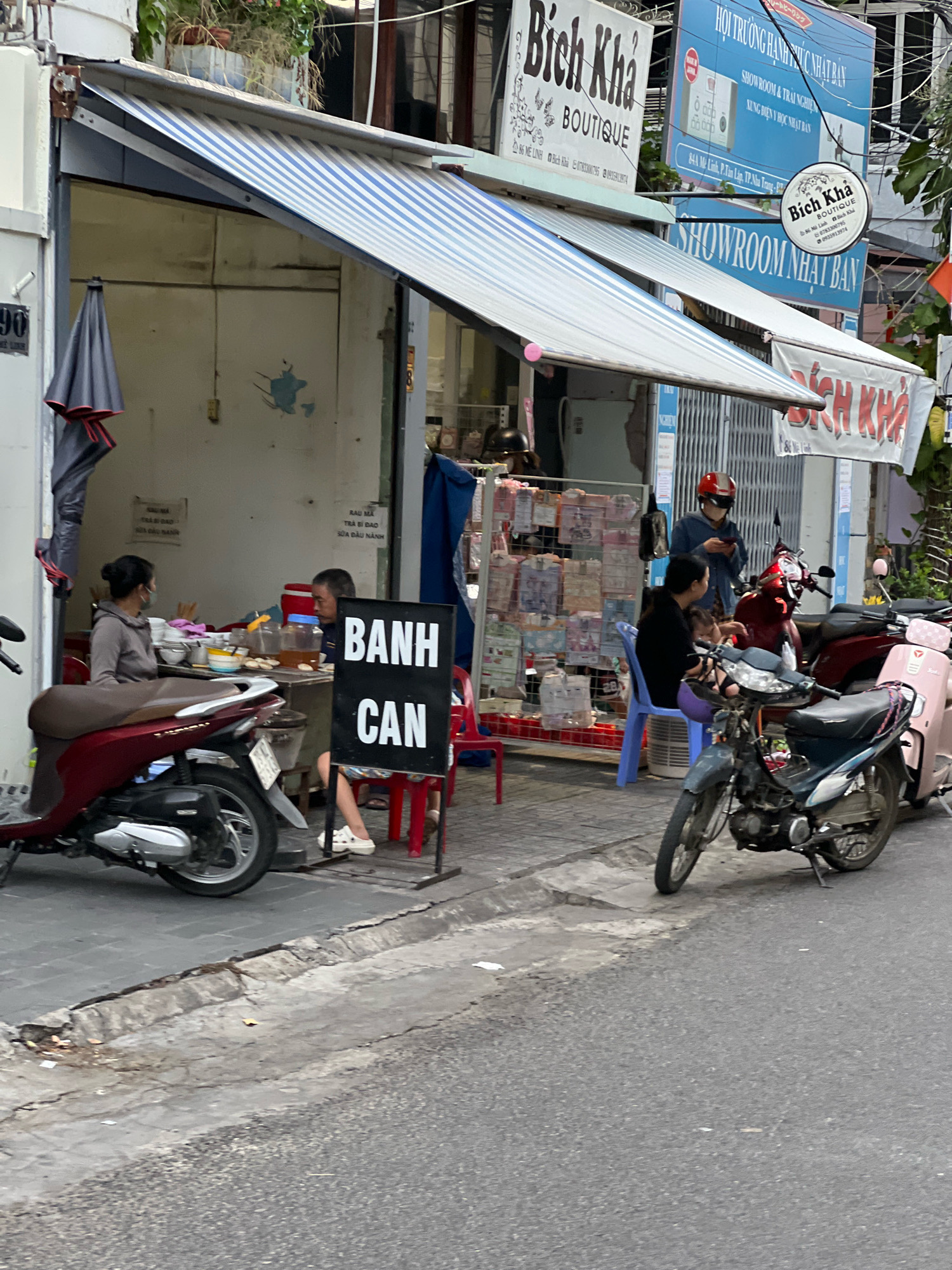 The image depicts a street scene in Nha Trang, Vietnam, likely in front of a shop. A woman with a mask on her face is seated at a table, facing away from the camera. A man is also seated at the table, looking towards the woman. The table is laden with bowls, utensils, and drinks. In front of the table is a black sign with white lettering that reads "BANH CAN". Parked motorcycles are visible on the left and right sides of the scene.
The shop appears to be an open-air establishment, with an awning extending over the seating area. Signs in Vietnamese are visible on the building, suggesting it is a commercial establishment. One sign, with the text "Bich Kha BOUTIQUE", indicates a boutique shop. Another sign, a circular one, also reads "Bich Kha Boutique". Further signs suggest it is a showroom for Japanese products, with text such as "SHOWROOM NHAT BAN". The environment is urban, with a paved street and sidewalk. Foliage is present in the background, suggesting it's a relatively green area despite the urban setting. The overall impression is a casual, everyday scene of street-side commerce and dining in Nha Trang.