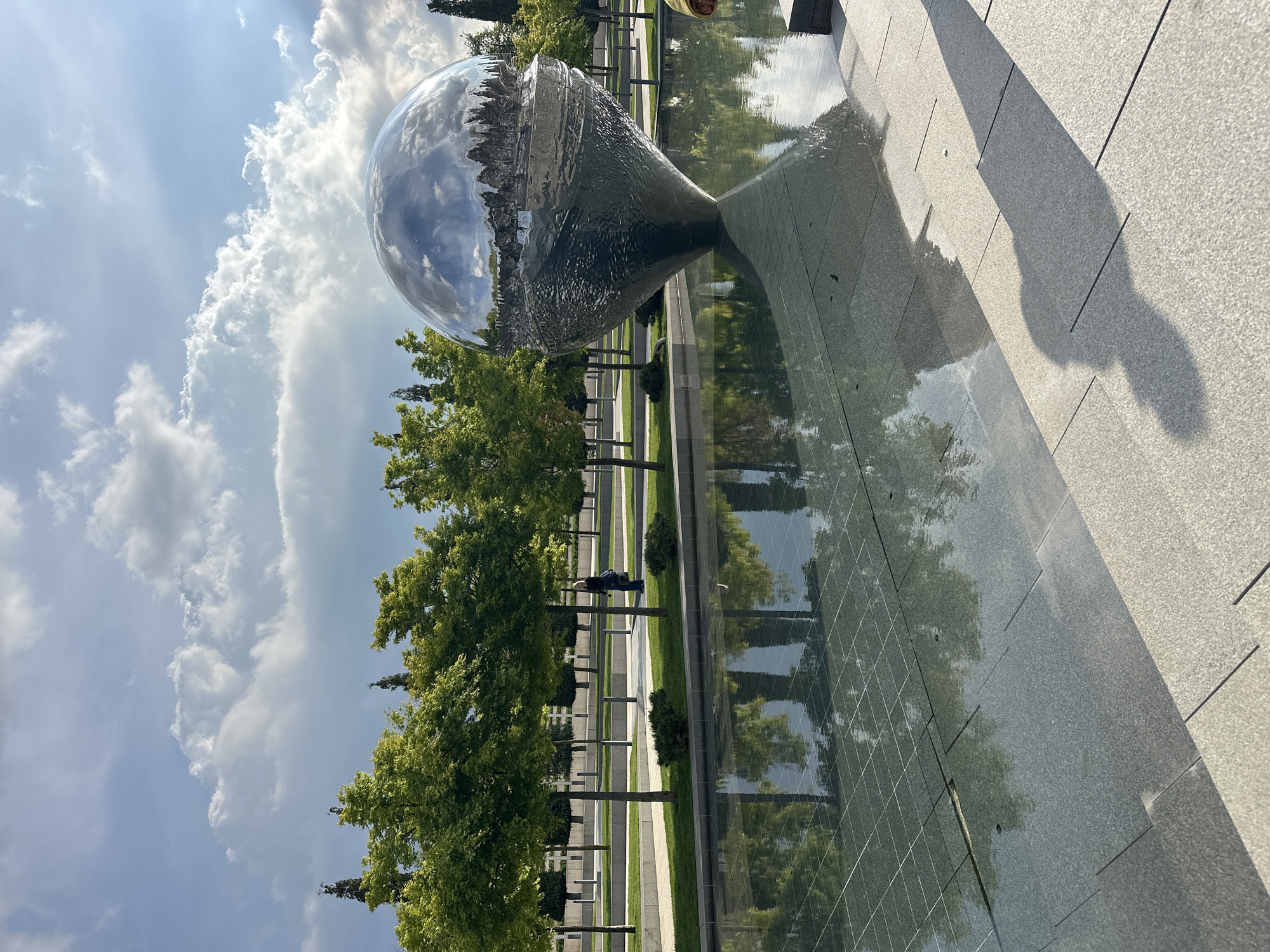 The image shows a mirrored art installation in the city of Krasnodar, Russia. The sculpture consists of a large, spherical object partially submerged in a shallow pool of water. The surface of the sphere reflects the surrounding environment, including trees, clouds, and the sky. A row of trees is positioned behind the pool. One person stands to the left, between the sculpture and the row of trees. The pool is surrounded by stone tiles with vertical lines running through them. The sky is partially covered with cumulus clouds.