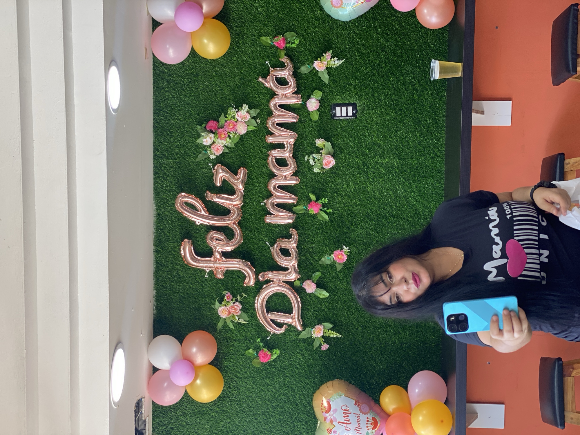 A woman is taking a selfie in front of a backdrop with the words "Feliz Dia Mama" (Happy Mother's Day) spelled out in rose gold balloons, decorated with small flowers. The backdrop is a green artificial turf, and there are bunches of balloons in pastel colors (pink, white, yellow, and orange) surrounding it. She's holding a blue cell phone. The location appears to be indoors, potentially a home or a decorated event space. The location is not explicitly identifiable as Juan Griego, Venezuela.