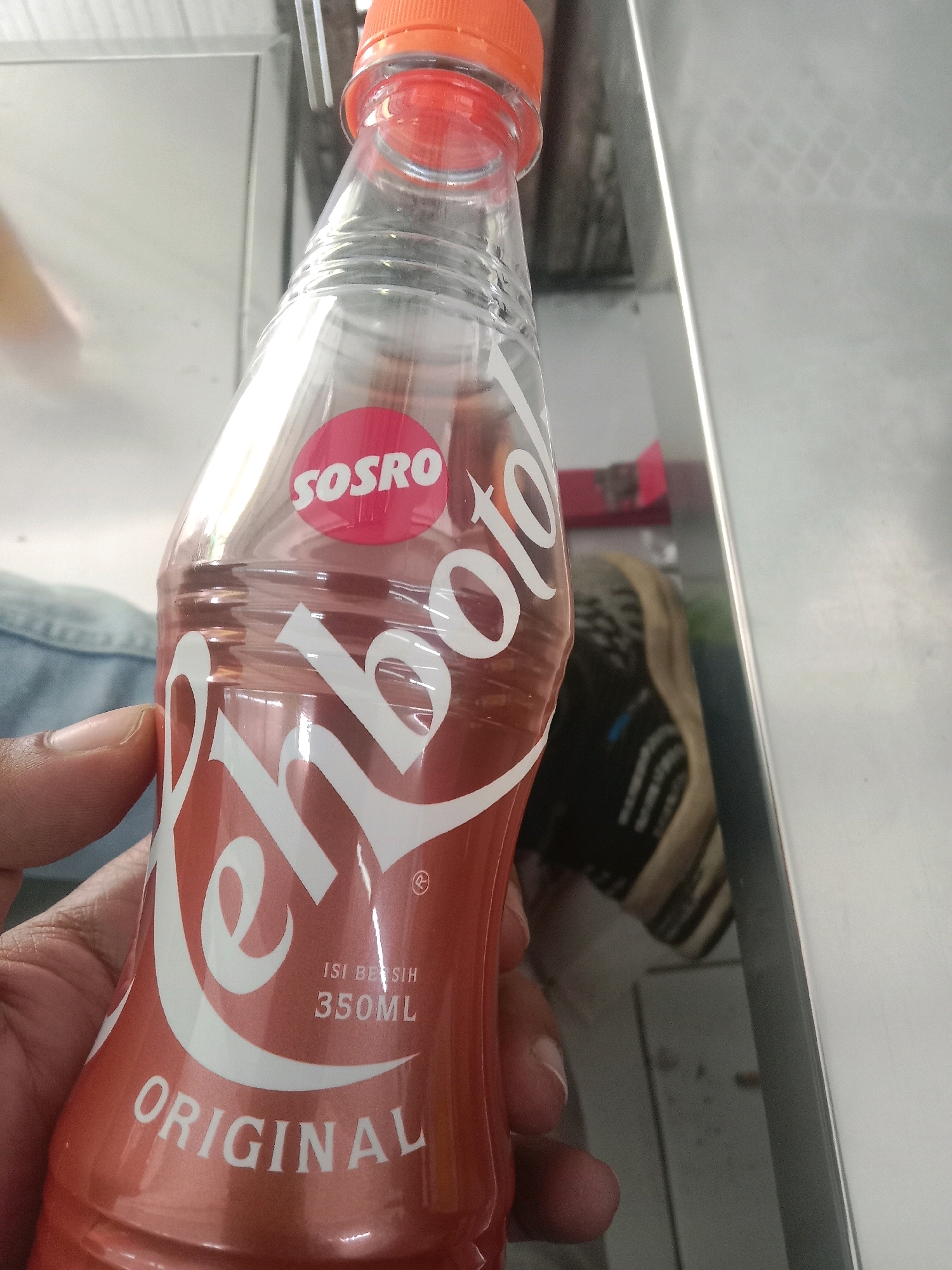 A hand is holding a bottle of Sosro Teh Botol Original. The bottle is made of clear plastic and contains a brown liquid. The label on the bottle is red and white and features the Sosro logo and the words "ORIGINAL" and "350ML". The background of the image is blurred and appears to be an indoor setting with metallic surfaces. The image was taken in the city of Kalikajar, Indonesia.