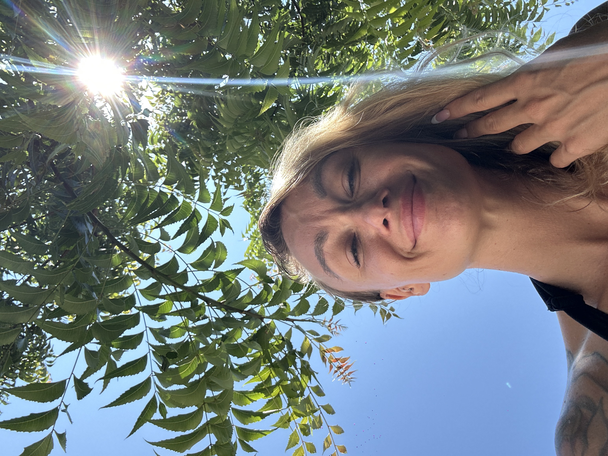 A low-angle, wide shot captures a young woman taking a selfie outdoors on a bright, sunny day in Mũi Né, Vietnam. The woman, with fair skin and blonde hair, is positioned in the lower half of the frame, smiling broadly. Her left eye is squinted, and her right eye is partially closed, suggesting she's looking directly into the sun, which is prominently featured in the upper right quadrant of the image. The sun's rays beam through the lush green foliage of a tree, creating a lens flare effect. The woman's right hand is raised to her head, touching her hair. She is wearing a black top. The background consists of a clear blue sky and the dense green leaves of the tree. A tattoo is visible on her left shoulder. The overall scene suggests a relaxed and joyful moment outdoors, possibly at a beach or park in Mũi Né.
