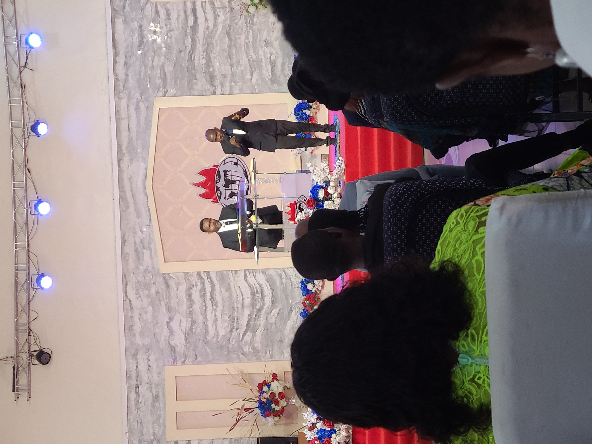 The image shows an indoor scene, likely a church or auditorium, with two men on a stage. The men are dressed in suits and appear to be speaking or presenting, possibly during a service or event. The stage has a backdrop with a logo or emblem and is decorated with flowers. The foreground shows audience members seated, with the backs of their heads and shoulders visible. There are lighting fixtures visible on the left side of the frame. The specific location in the city of Unknown, Unknown is not discernible from the image.