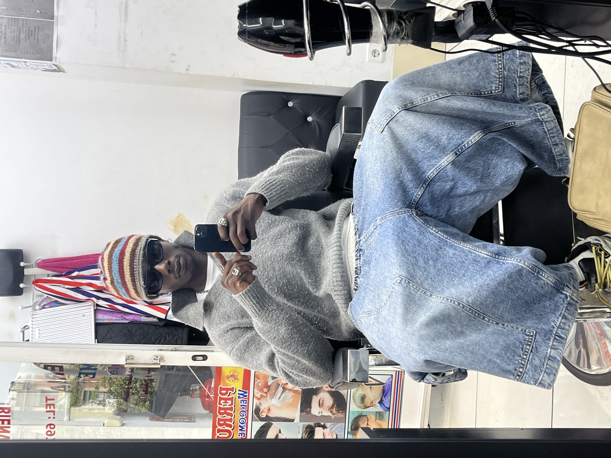 A person wearing a multicolored beanie, sunglasses, a gray sweater, and baggy blue jeans is sitting in a barber chair, taking a photo with a smartphone in a barbershop. The reflection in the mirror reveals details of the barbershop, including the advertisement for barbershop services in the window. The location is Barcelona, Spain, as implied by the barbershop advertisement in Catalan.