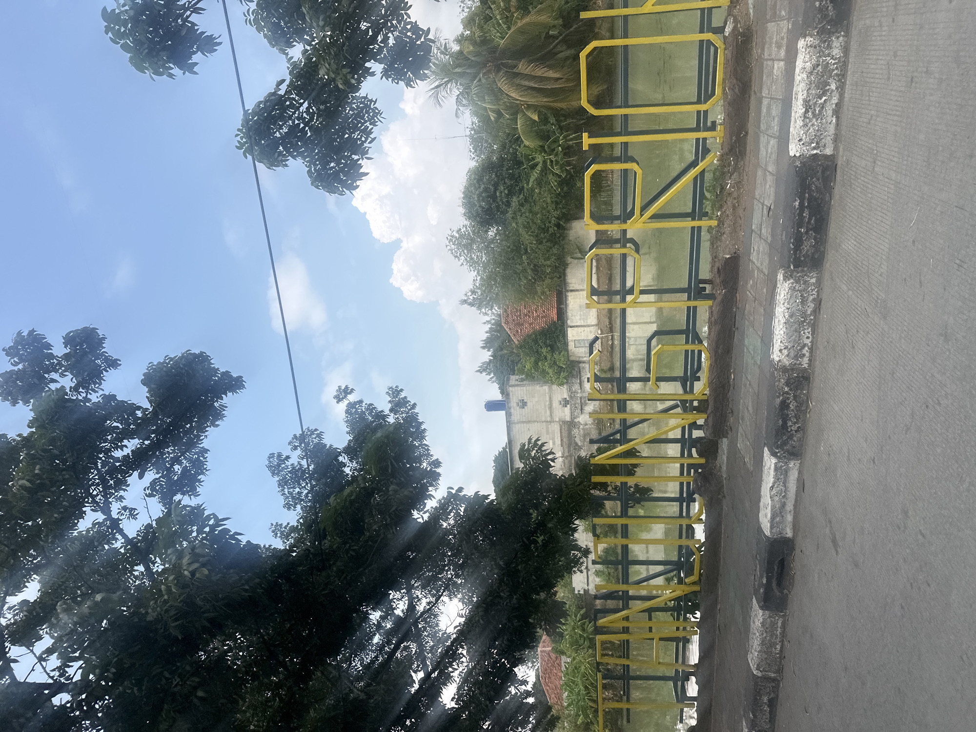 The image shows a street scene in North Jakarta, Indonesia. The background includes trees, a building, and a body of water, possibly a canal or river. Yellow lettering spells out "SUNDANG" on a barrier along the edge of the water. To the right of the lettering is a sidewalk with black and white markings and a paved road. The sky is visible in the upper left corner and includes trees, clouds, and a blue sky. The image appears to have been taken from inside a vehicle.
