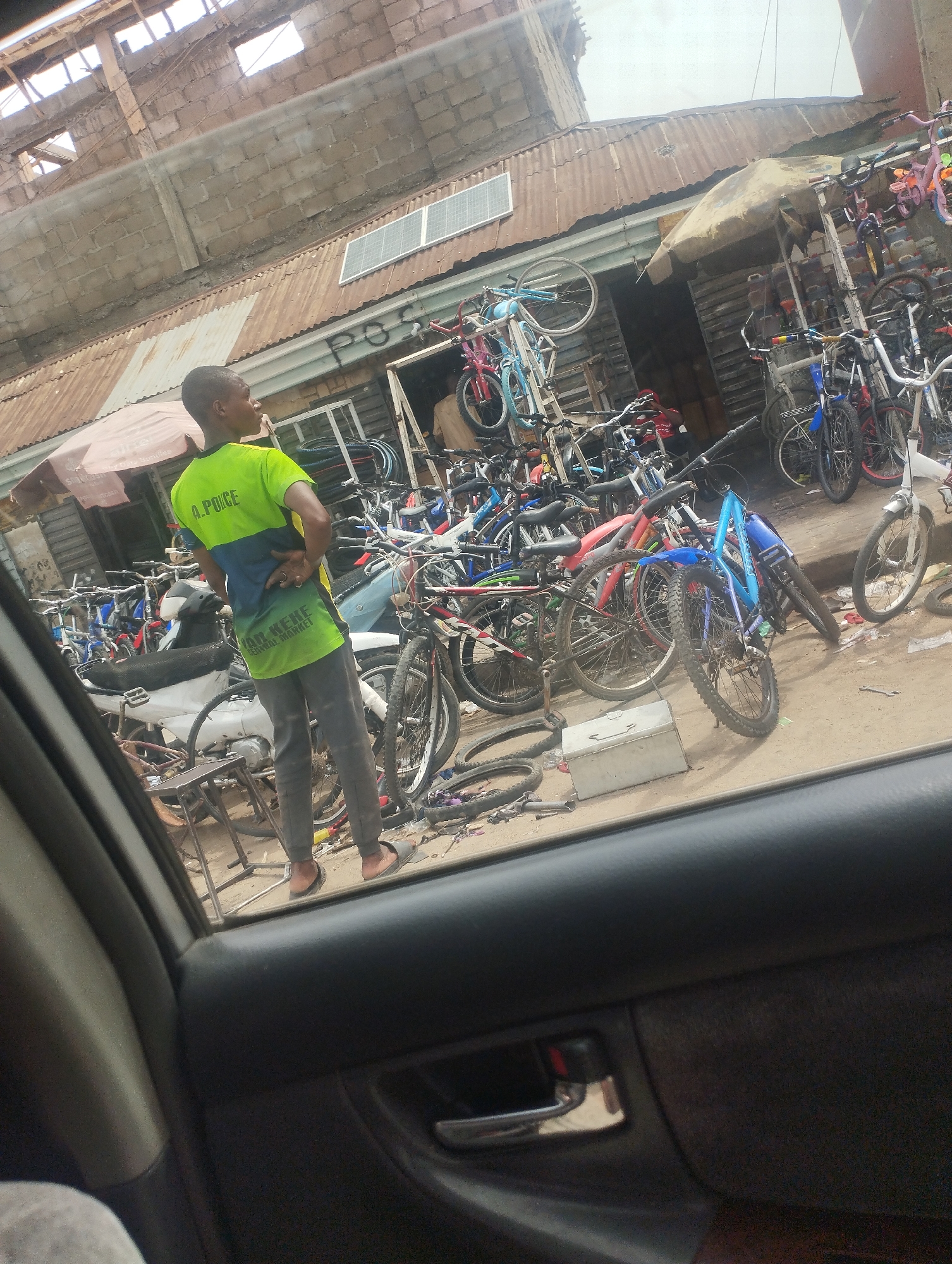 Captured from inside a car, the image shows a bustling scene likely in Bauchi, Nigeria, indicated by the writing on the t-shirt of the man in the shot. The focus is on a local bicycle shop. The shop is overflowing with numerous bicycles of various sizes and colors, partially sheltered by a makeshift awning. A man wearing a green and blue shirt stands with his back to the viewer, seemingly observing the shop. The shop is crowded with bikes and motorcycle. The architecture indicates a commercial area with buildings of concrete and corrugated metal.