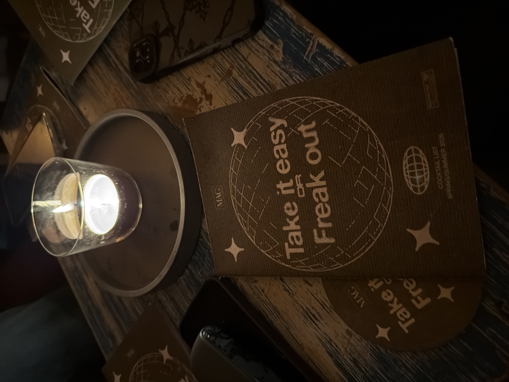 The image presents a dimly lit indoor scene, likely a bar or restaurant in Milan, Italy, during the evening or night. The primary subject is a lit candle, housed within a clear glass cylinder and placed on a dark, circular base. Its soft glow illuminates the immediate surroundings, creating an intimate atmosphere.
The candle rests on a rustic wooden table, which features a distressed finish with visible grain and hints of blue and white paint, suggesting wear and character. On the table, a dark brown coaster or menu is prominently displayed. It features the bold white text: "Take it easy OR Freak out," set within a stylized globe illustration. Below this, the text "COCKTAIL LIST" and "SPRING/SUMMER 2026" is visible, along with a small logo (possibly "Sprecto" as per OCR, though indistinct) and the letters "MAG." A second, partially obscured, rounded coaster or menu below it shows similar design elements and the partial text "Take it Fre." Another menu is visible in the upper left corner with "MAG" and "Take it."
A smartphone in a dark, patterned case is also partially visible on the table. The overall scene evokes a relaxed, cozy setting, typical of a casual social gathering or a quiet moment, potentially while enjoying or waiting for drinks.