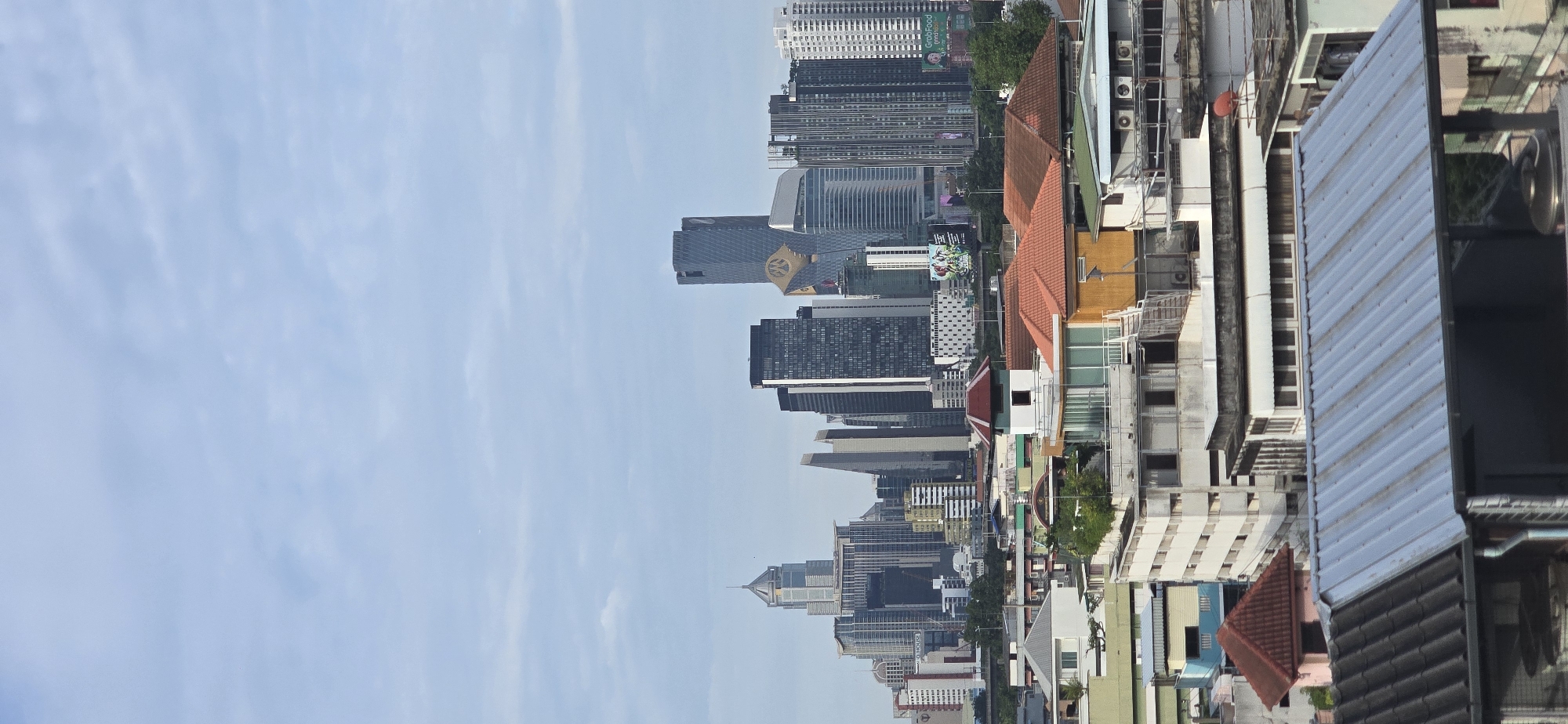 The vertical image captures a cityscape dominated by a hazy sky and a dense collection of buildings in Bangkok, Thailand. In the foreground, several multi-story residential or commercial structures are visible, characterized by flat roofs, some with corrugated metal sheeting, and varying architectural styles. These buildings show signs of age and wear, with visible pipes, air conditioning units, and some overgrown vegetation on rooftops.
Further back, a more modern skyline emerges, featuring numerous skyscrapers of varying heights and designs. These taller buildings are more contemporary, with reflective glass facades and angular shapes. One prominent skyscraper in the middle ground displays a distinctive golden emblem or logo on its upper section. Signs and advertisements, including a "GrabFood" advertisement, are visible on some of the buildings in the mid-ground and background.
The overall impression is one of a bustling urban environment with a blend of older and newer construction, set against a cloudy or hazy sky typical of a tropical climate. No people are clearly discernible in the image, and no specific actions or interactions are evident. The scene appears to be captured from an elevated viewpoint, looking across the city.