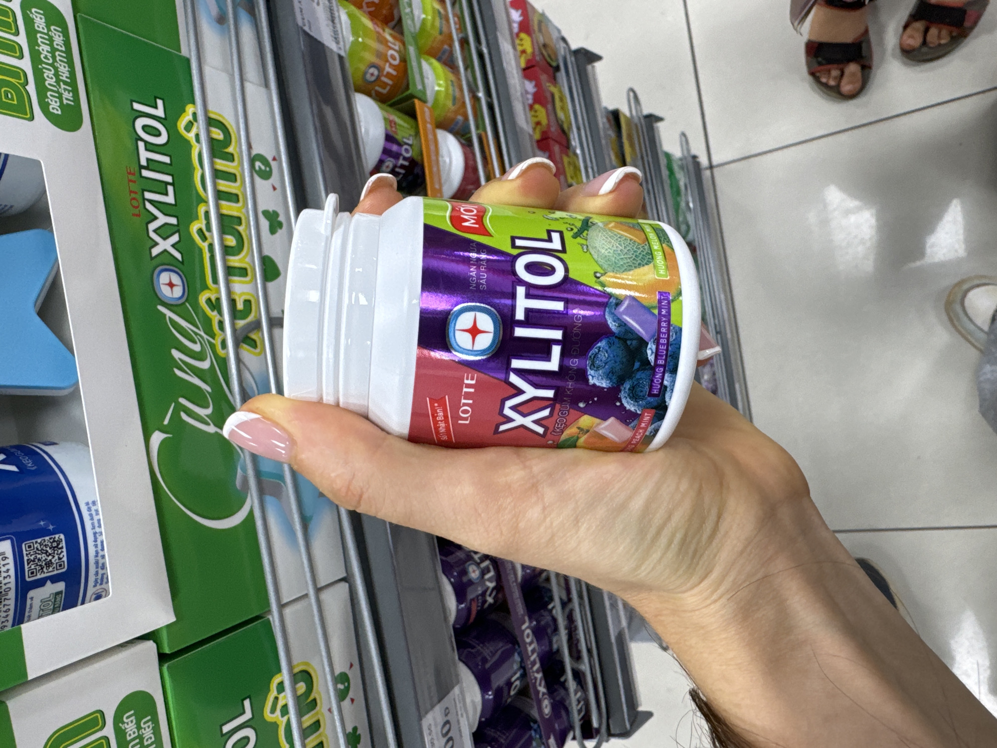 A hand with manicured fingernails holds a container of Lotte Xylitol gum in a supermarket aisle. The container, primarily white and purple, features a blueberry and melon flavor design. Several similar containers of gum line the shelves behind. The labels are written in Vietnamese and advertise the product's cavity prevention benefits. A green box of Xylitol gum is also in the lower left. The setting appears to be a retail environment, likely a supermarket or convenience store in Phu Thuy Ward, Vietnam. The background shows the tiled floor. Parts of two pairs of feet with sandals are visible.