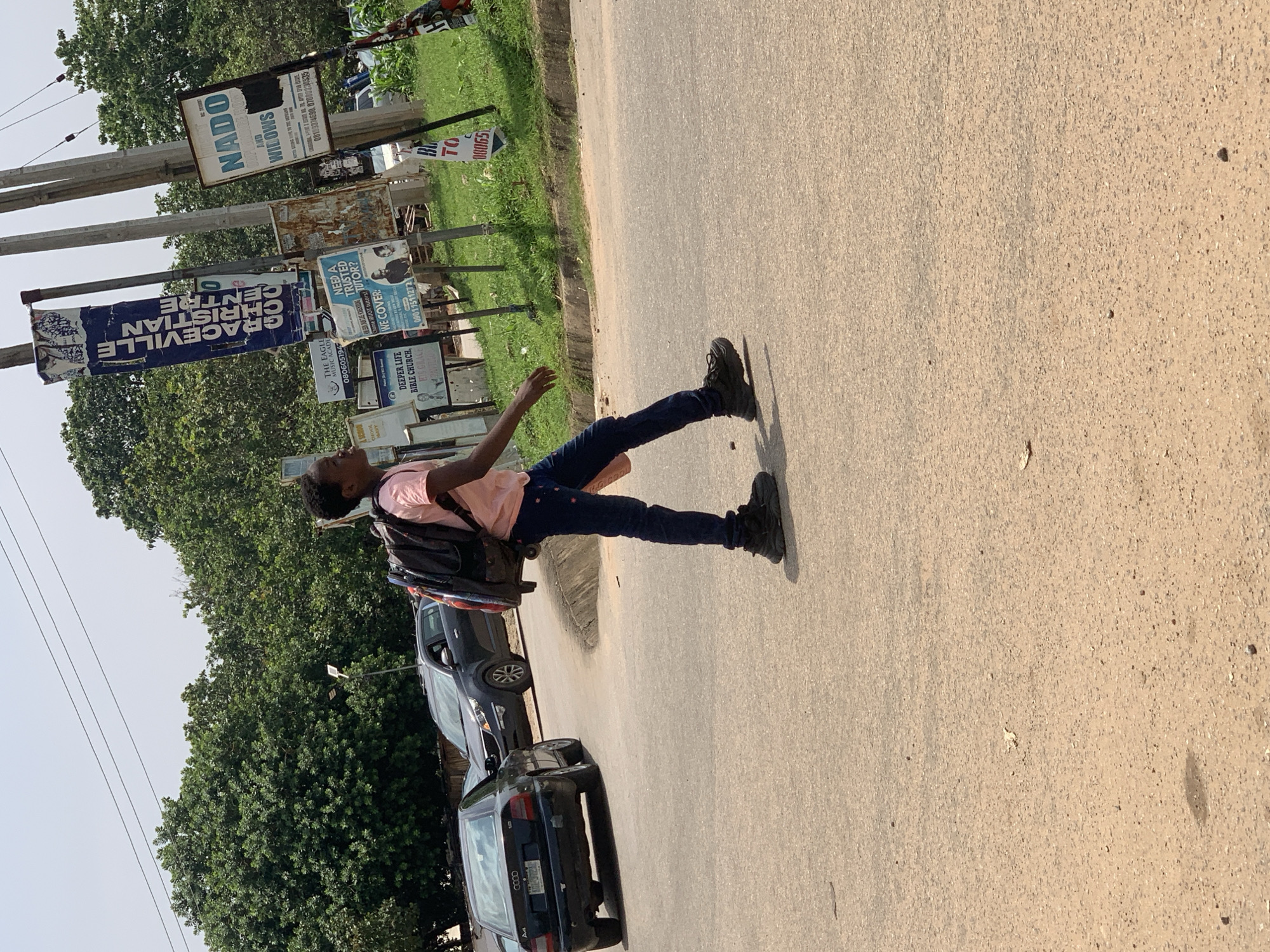 The image depicts a sunny daytime scene on a road in Abuja, Nigeria. A young person wearing a pink t-shirt, dark blue jeans, and black sneakers is walking across the road from right to left, carrying a large black backpack. The person is in the foreground, with their back mostly to the camera, facing away from a dark blue Audi sedan and a dark grey SUV parked or driving on the road behind them. The road surface is asphalt.
In the background, there is a grassy area with several utility poles adorned with various signs and banners. One prominent blue banner reads "GRACEVILLE CHRISTIAN CENTRE". Other signs include one for "NADO AND WILLOWS", and several smaller, weathered signs. Lush green trees and foliage line the background, suggesting a suburban or semi-urban environment. Overhead, power lines are visible against a pale sky. The overall impression is of a casual street scene in a city.