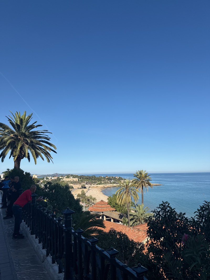The image shows a sunny coastal scene in Tarragona, Spain, featuring a viewpoint overlooking a beach. A group of people, including a man in a red shirt leaning over a black railing, are observing the view. Palm trees frame the scene, with buildings featuring orange-tiled roofs visible. The Mediterranean Sea stretches to the horizon under a clear blue sky.