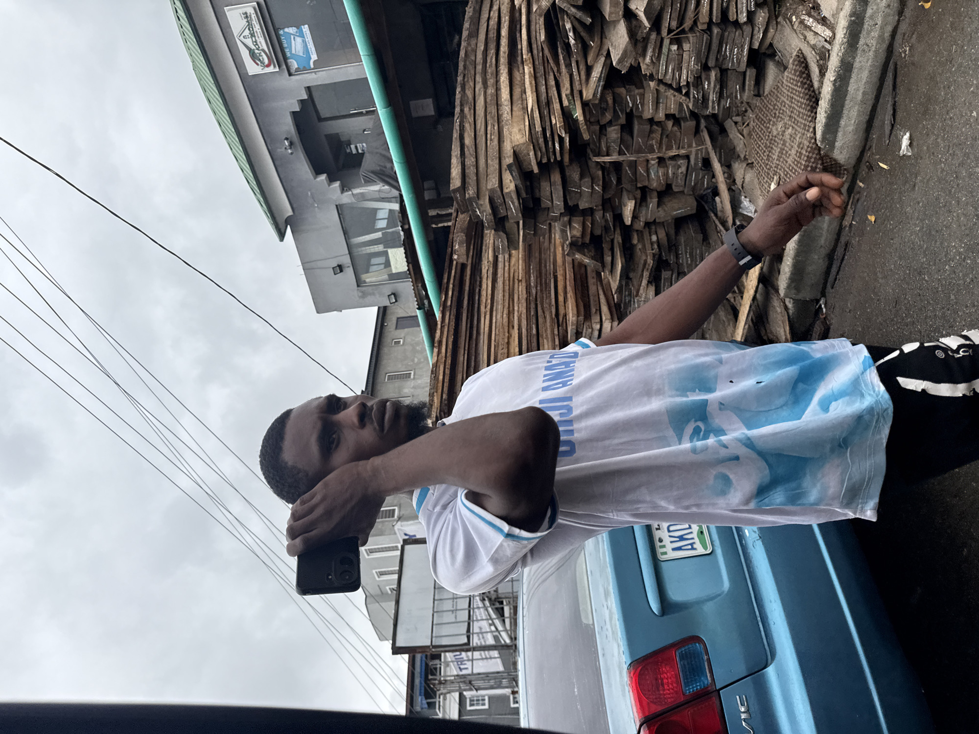 The image depicts a young Black man standing on a street in Ado, Nigeria, holding a smartphone to his ear as if on a call. He is wearing a white t-shirt with blue graphics and black athletic pants. To his right, a large stack of wooden planks is piled high. Behind him, a light blue sedan is parked. The background shows a building with a sign that appears to be for a business. Overhead, utility wires crisscross the cloudy sky. The overall environment suggests an outdoor commercial area, possibly a marketplace or a construction supply yard.