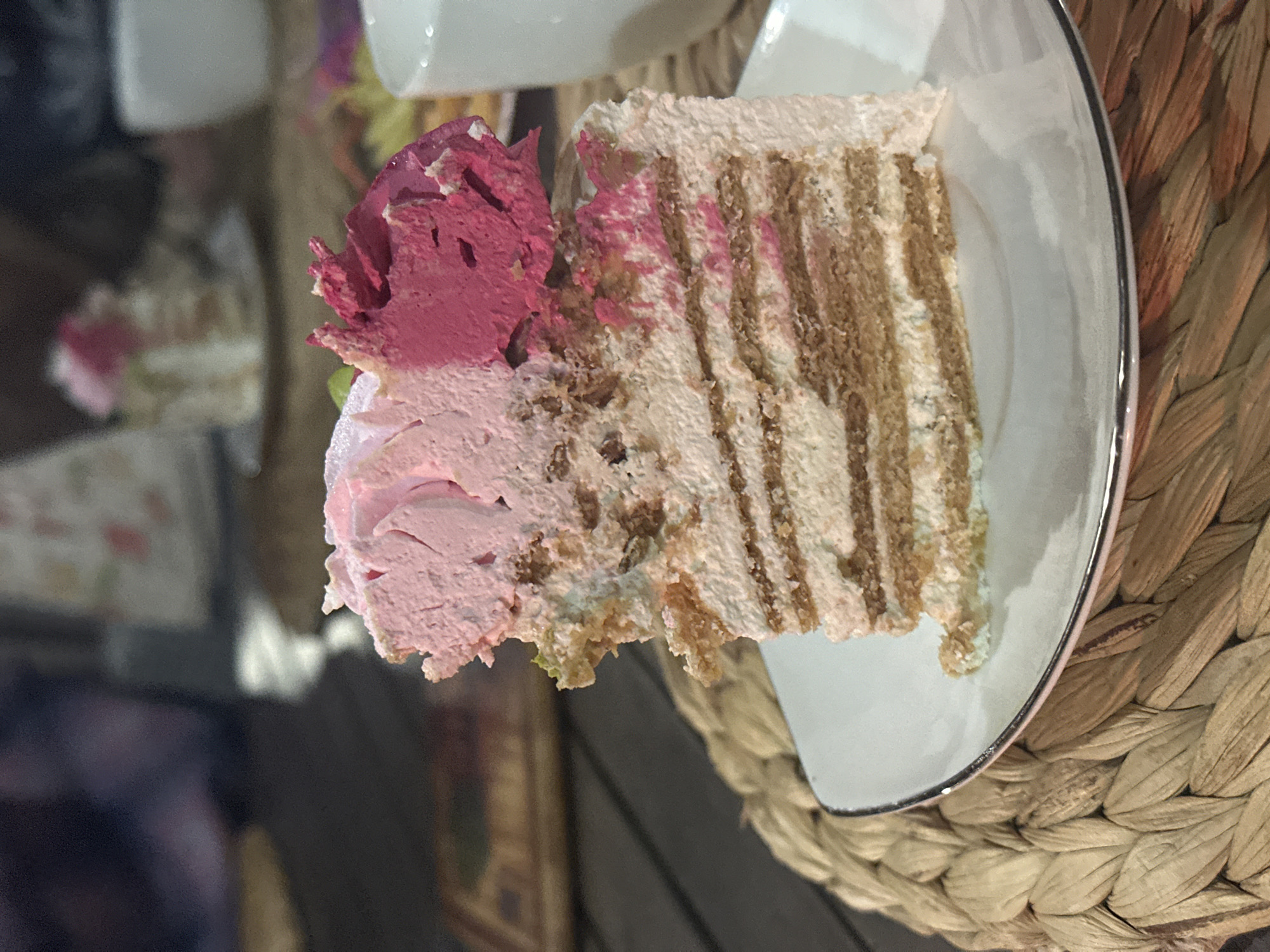 A slice of layer cake sits on a white plate with a silver rim, placed on a woven mat. The cake features layered cake and frosting, with pink and magenta frosting dollops on top. The background includes blurred objects and surfaces. The location and relation to the city of Aviator, Russia is indeterminable.