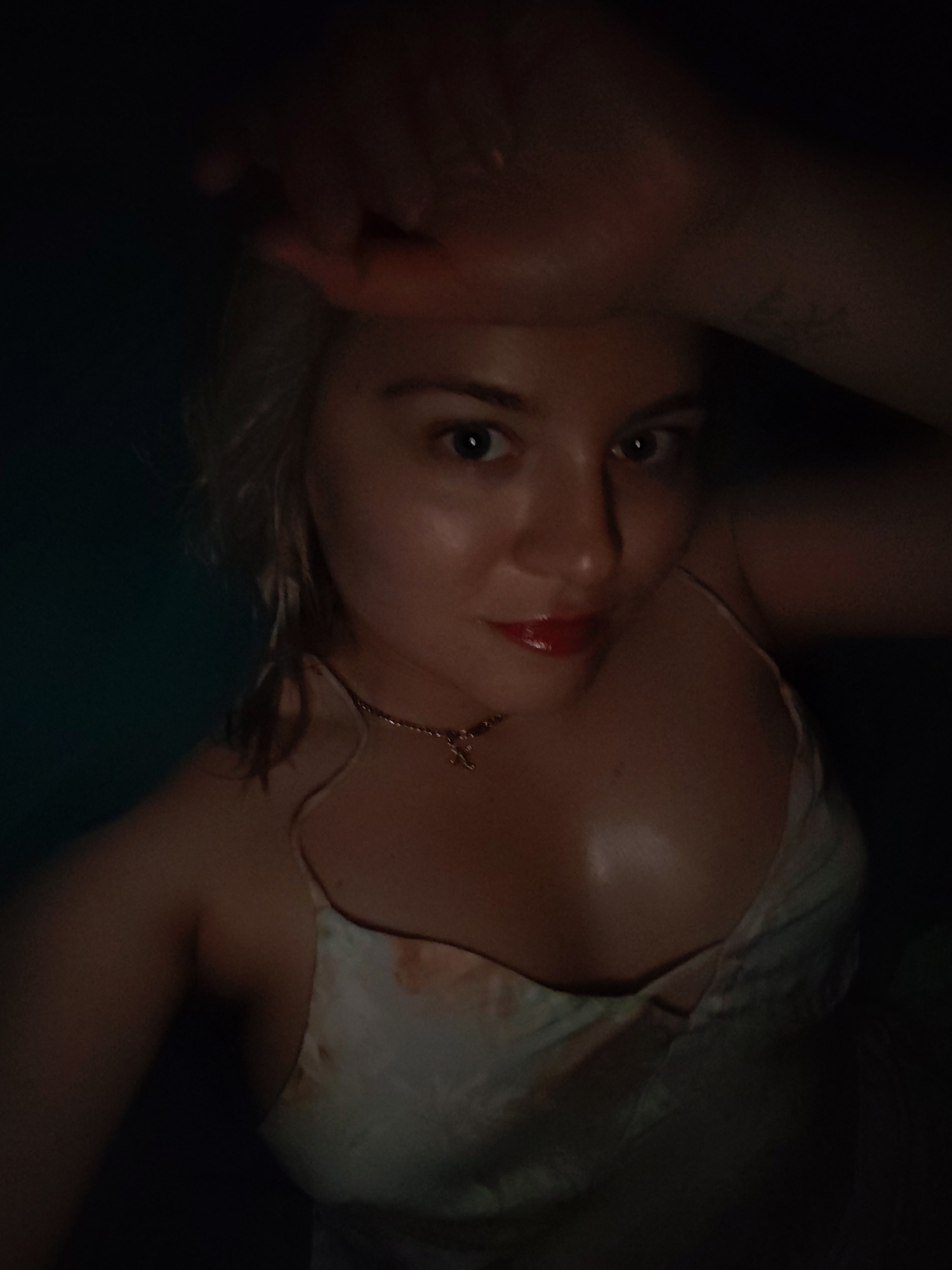 A woman is shown in a selfie-style image with her hand raised near her forehead, partially obscuring the upper left of the frame. She has light skin, blonde hair, and is wearing red lipstick. A gold necklace is visible around her neck. She is wearing a light-colored, patterned dress. The background is dark and undifferentiated. There is a tattoo on her right forearm. There is no information to suggest that this image was taken in Izhevsk, Russia.