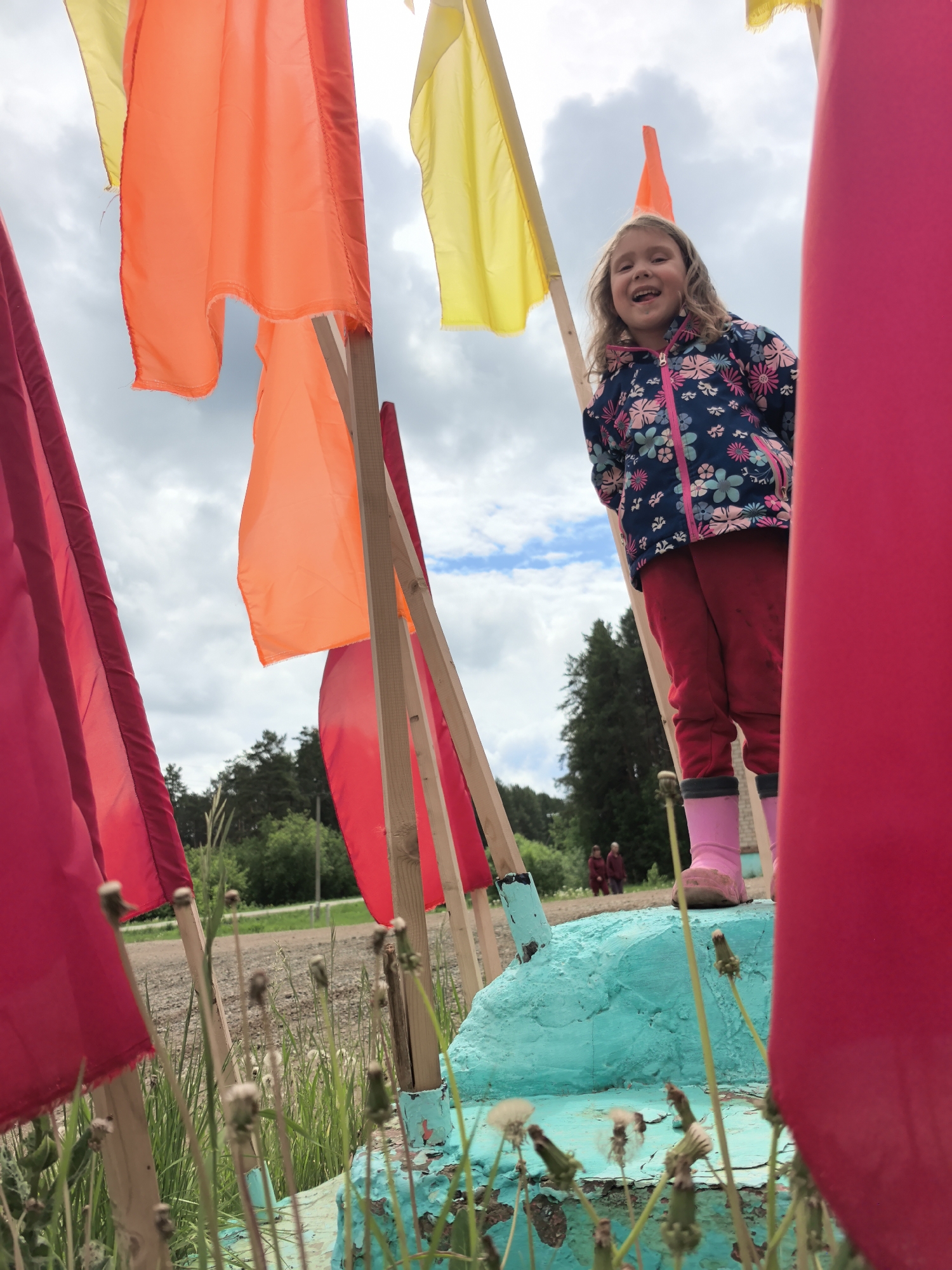 A young girl stands on a turquoise concrete structure surrounded by colorful flags in a field. The flags are red, orange, and yellow and are attached to wooden poles. The girl wears a floral jacket, red pants, and pink boots. She has blonde hair and is smiling. The background shows a cloudy sky, trees, and a dirt road with figures in the distance. The location appears to be a grassy area with dandelions and other vegetation. The image appears to have been taken in Shabury, Russia.