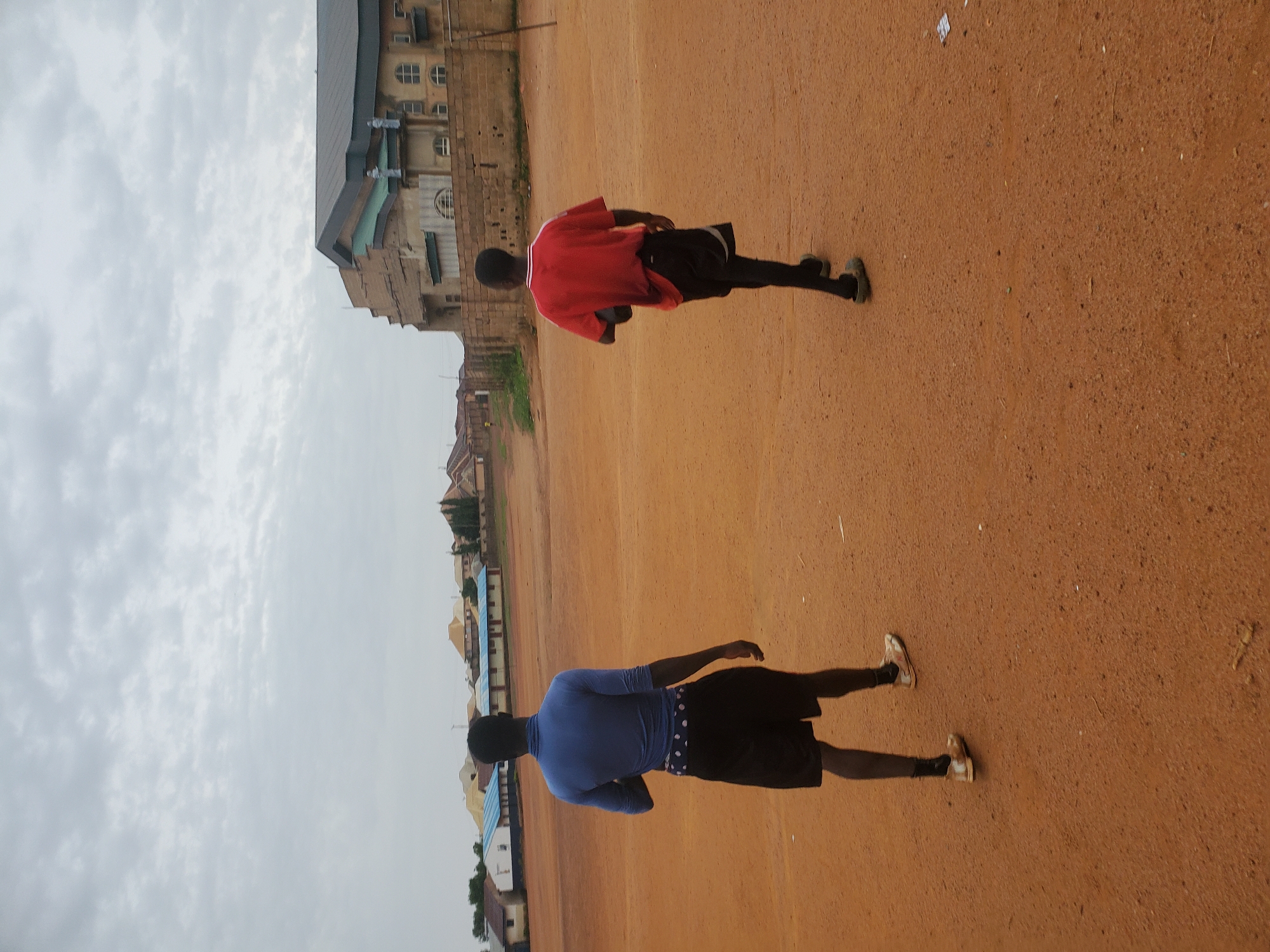 The image depicts two people standing on a large expanse of red dirt, likely a playing field. The person in the foreground is wearing a blue long-sleeved shirt and black shorts. The person further away is wearing a red shirt and black shorts. Both individuals are facing away from the camera. In the background, there is a building with a multi-tiered roof and several windows. Beyond that, there are houses and trees. The sky is overcast. Based on the architectural style and general environment, the location is likely in Jos, Nigeria.