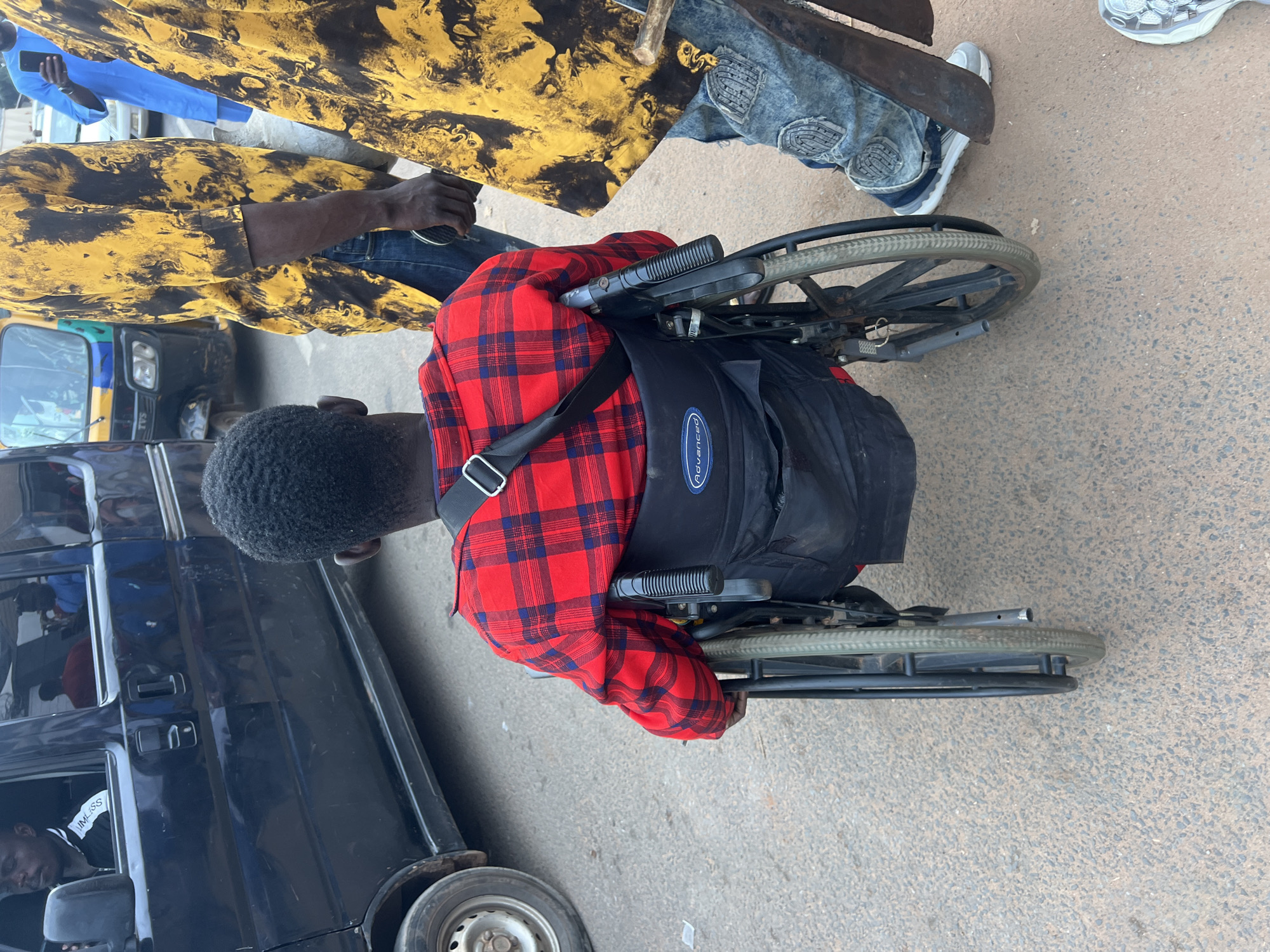 In Nkpor, Nigeria, a man with dark skin and short, dark, curly hair sits in a black wheelchair, viewed from behind. He is wearing a red and blue plaid long-sleeved shirt. The wheelchair has black upholstery and large black wheels with metal spokes. A black strap is visible across the back of his shirt. Next to him, on the right, another person stands, wearing a yellow and black patterned shirt and blue jeans. The person's right hand is visible, holding an object that appears to be a machete. In the background, a black minivan is parked. The scene appears to be on a paved road with visible traffic.