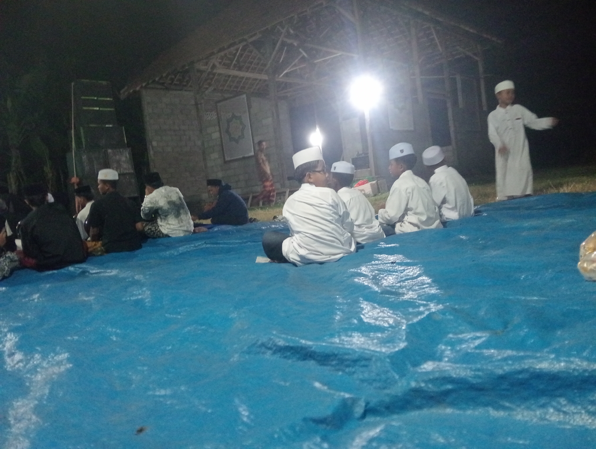 In Bloro, Indonesia, a group of young boys and men, mostly wearing white Islamic attire and peci caps, are gathered outdoors at night. They are seated on a large blue tarp, facing a simple, open-sided structure with a thatched roof. A bright light source illuminates the scene from within the structure, casting long shadows. The background shows a brick wall, some vegetation, and a tall, dark structure that appears to be some sort of scaffolding or equipment. One boy in a white robe is standing and gesturing. The overall atmosphere suggests a religious gathering or educational event.
