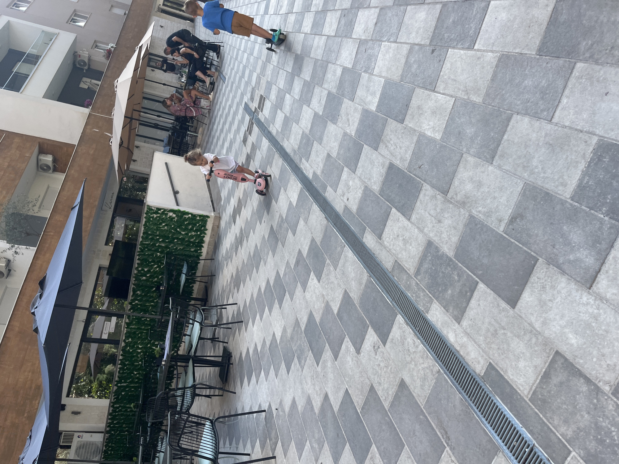 In the city of Budva, Montenegro, a paved outdoor patio area features a drainage grate running diagonally across the foreground. Two young children are riding scooters on the patio. A girl, in a white shirt and pink shorts, rides a pink scooter. A boy, in a blue t-shirt and tan shorts, rides a green scooter. In the background, several people are seated at tables under umbrellas outside a building with large windows. The building appears to be a restaurant or cafe. Outdoor seating with tables and chairs is visible in front of the building, some covered by dark blue umbrellas. The patio is paved with a repeating pattern of light and dark grey rectangular tiles. Green hedges are visible behind the tables and chairs.