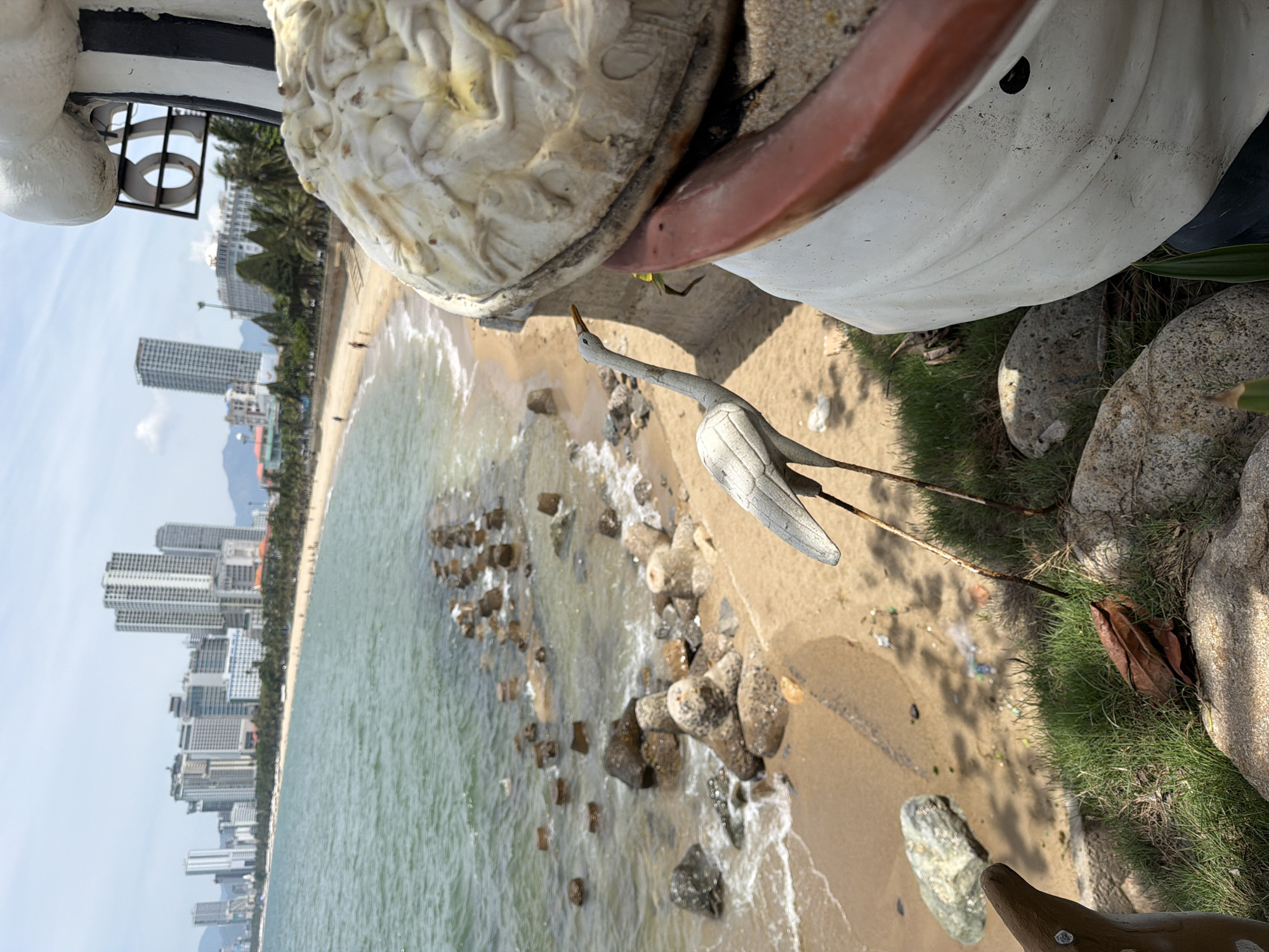 This image depicts a coastal scene in Nha Trang Ward, Vietnam. In the foreground, a statue of a white bird with a long neck and yellow beak stands on thin legs, positioned on a grassy area with rocks. To the right, a large, rounded white sculpture with textured detailing and a reddish-brown rim partially obscures the view of the bird and the background. The midground reveals a sandy beach with a gentle wave line and scattered rocks and breakwaters extending into the sea. The water is a greenish-blue, with visible ripples and waves. In the background, a long stretch of coastline is visible, lined with palm trees and numerous tall, modern buildings characteristic of a city skyline. The sky is bright and appears partly cloudy. Several individuals are walking along the beach in the distance. The overall impression is that of a city beach.