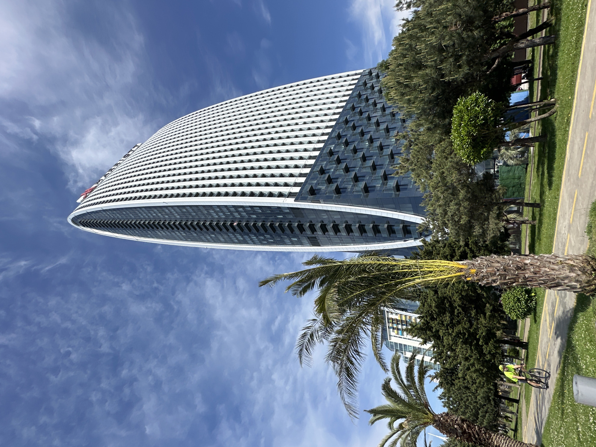 The image captures a unique architectural design in Batumi, Georgia. A tall, elliptical building dominates the view, featuring a white facade with geometric windows on one side and a darker, glass-paneled structure on the other. The building's curvature emphasizes its modern style.
To the right, a mix of lush greenery, including palm trees and other trees, creates a natural contrast with the building's sharp lines. A paved path runs along the side, and a cyclist wearing a neon yellow shirt can be seen riding in the distance, adding a sense of human activity to the scene.
The sky above is bright blue with scattered white clouds, providing a vibrant backdrop. The overall composition suggests a blend of urban development and natural beauty, typical of Batumi's coastal landscape.