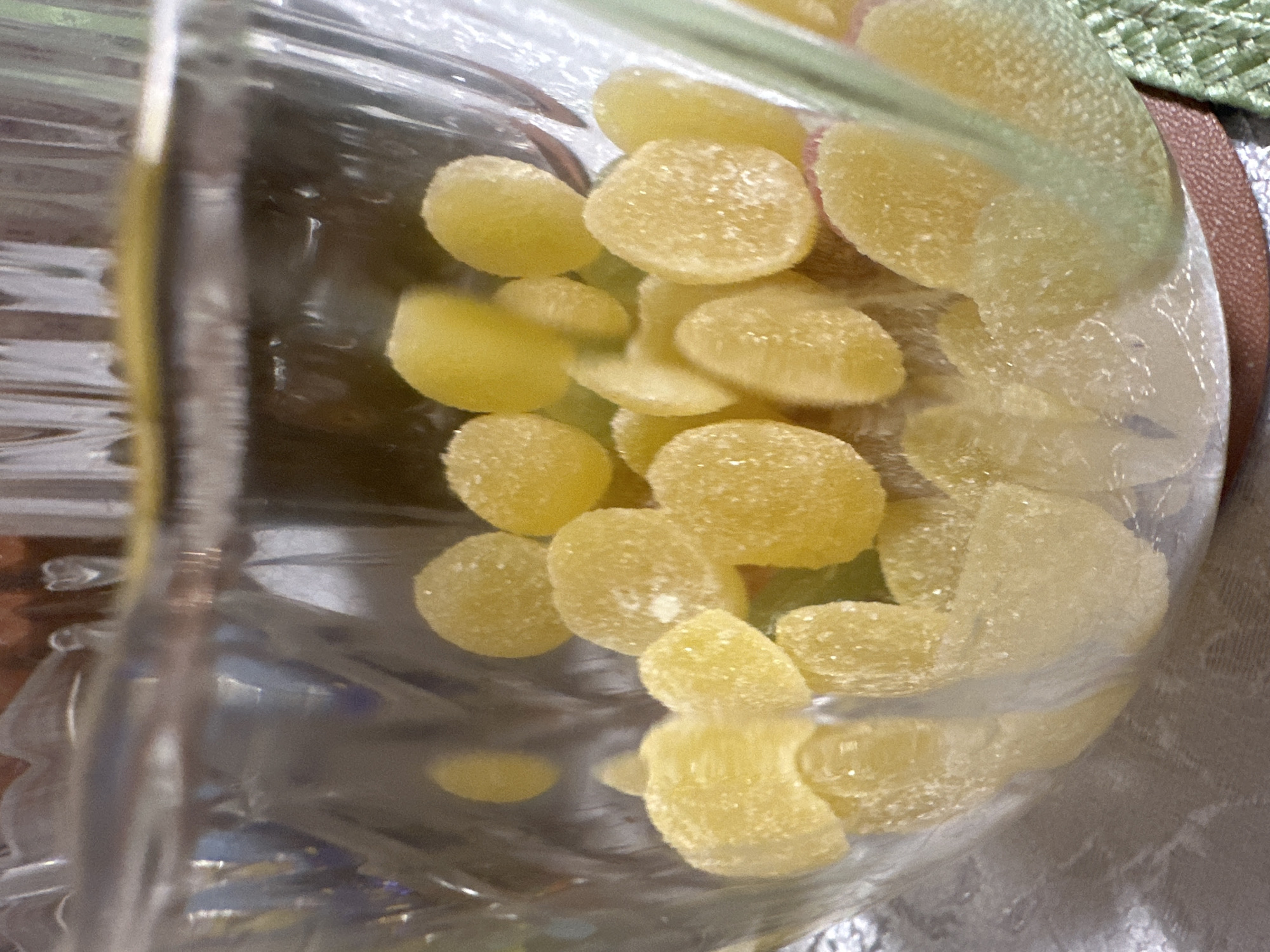 The image depicts a clear, likely plastic, container filled with yellow, round, sugar-coated candies. The container appears partially full, with the candies clustered together. The background shows a blurred surface, potentially a table, with a hint of green and brown. The image is taken indoors with natural lighting. There is no verifiable connection to Vorotynsk, Russia in the image. The description contains no information on people, subjects, actions, interactions, or environment beyond the visual objects present in the image.