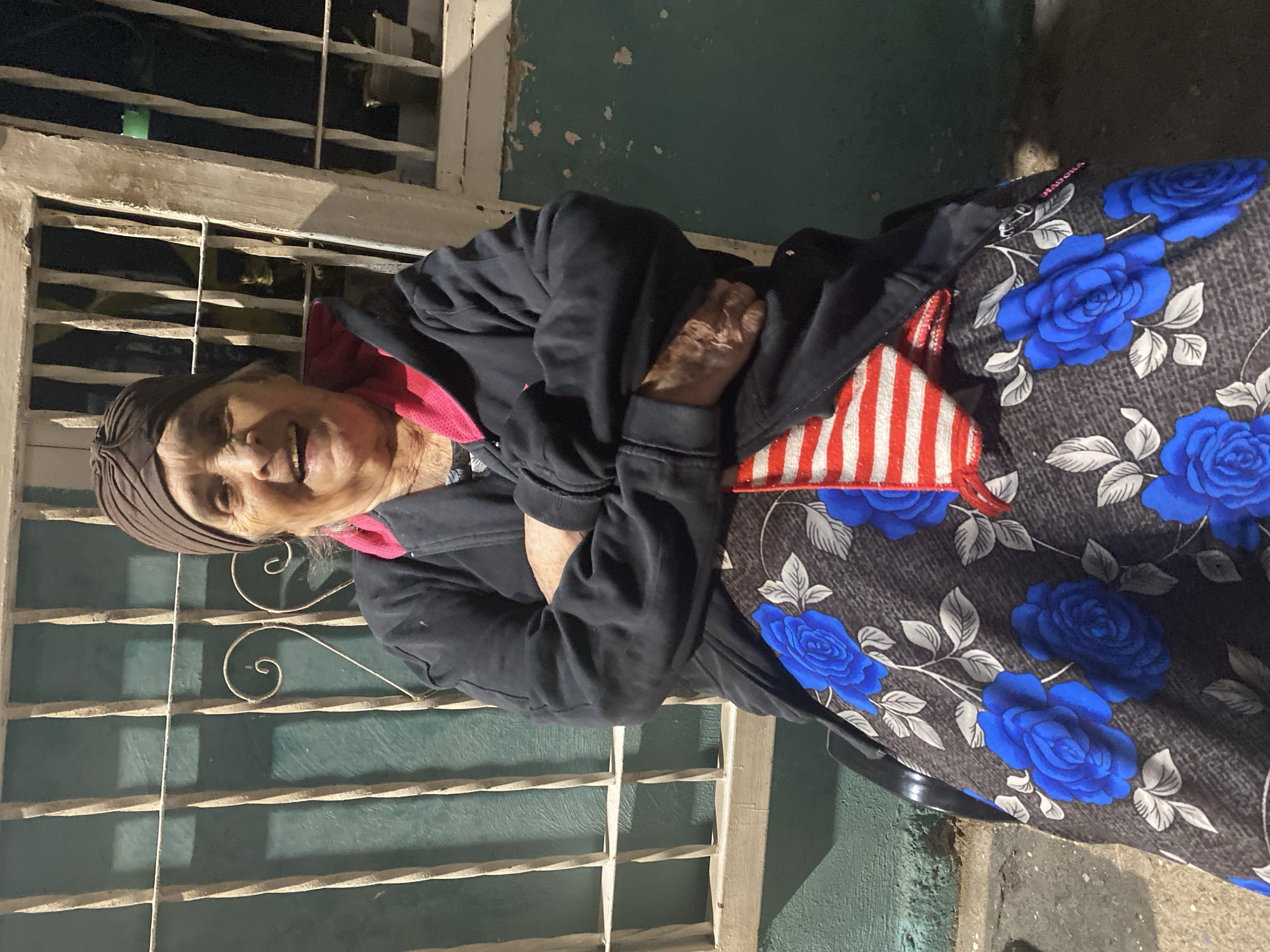 In El Cambur, Venezuela, an elderly woman with grey hair, wearing a brown headscarf and a black jacket over a pink garment, sits with her arms crossed. She is adorned with a dark skirt featuring a blue rose floral pattern. A red and white striped fabric is tucked into her skirt. The background features a light-colored, barred window or gate against a dark green wall. The scene appears to be outdoors at night, with artificial lighting casting shadows on the wall and ground.