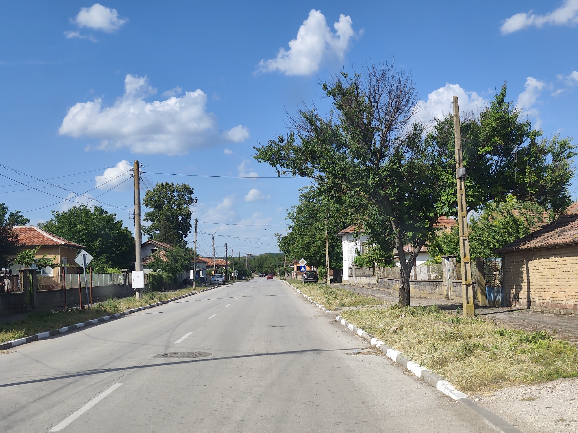 The image shows a straight, paved road running through a village, likely in Balgarene, Bulgaria, based on the context. The road has white lane markings and a manhole cover is visible on the asphalt. On both sides of the road, there are houses with red-tiled roofs, some with brick walls and others with painted facades. Trees and vegetation are present along the roadside, with some dry grass and weeds in the foreground. Utility poles with wires are interspersed throughout the scene. A few cars are visible on the road and parked in front of some of the houses. The sky is blue with scattered white clouds. The scene appears to be a rural or suburban setting on a clear day.
